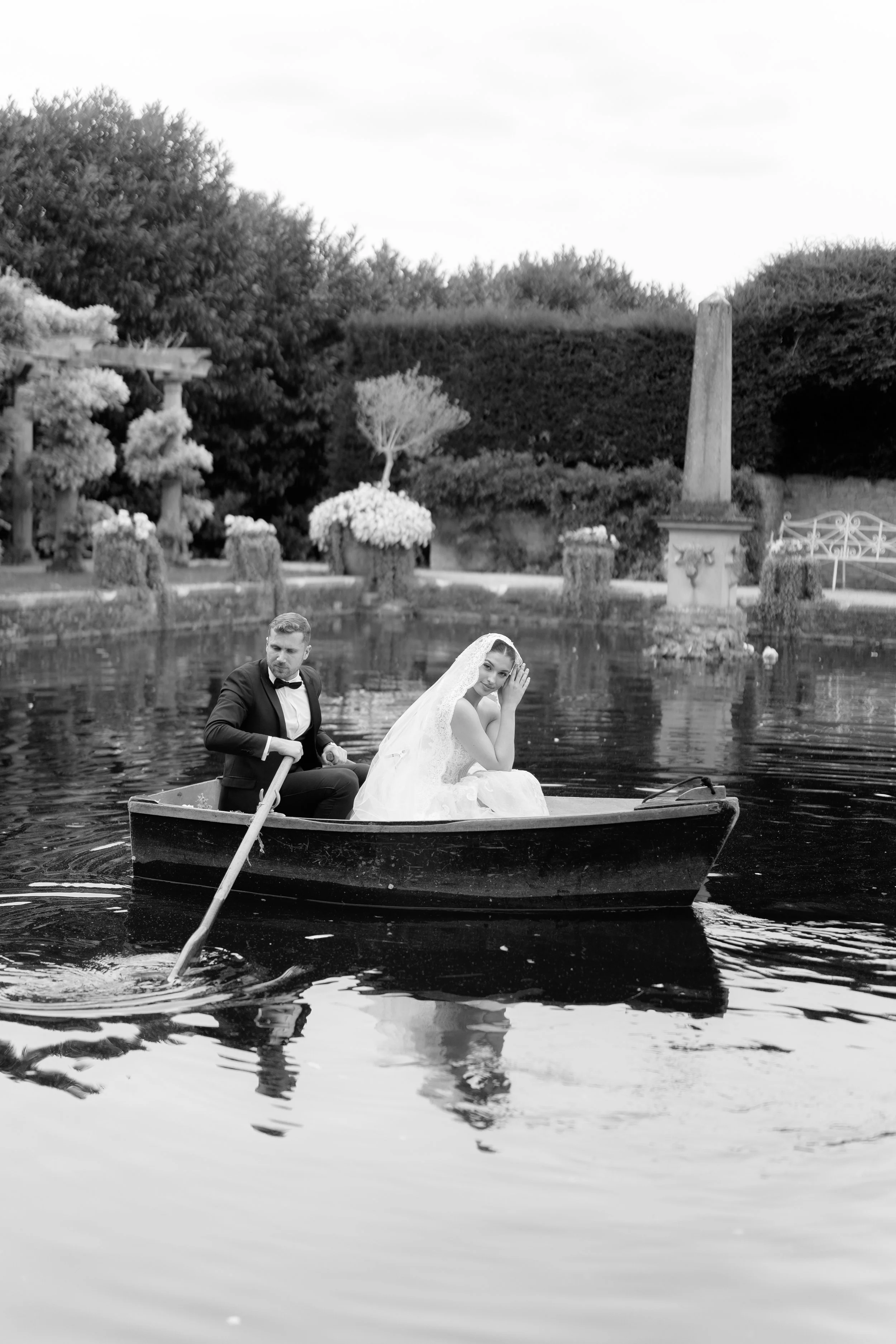 Black and white photo of a bride and groom in a small boat on a pond in a garden. The groom is rowing while the bride is sitting, wearing a wedding dress and veil, touching her face gently.