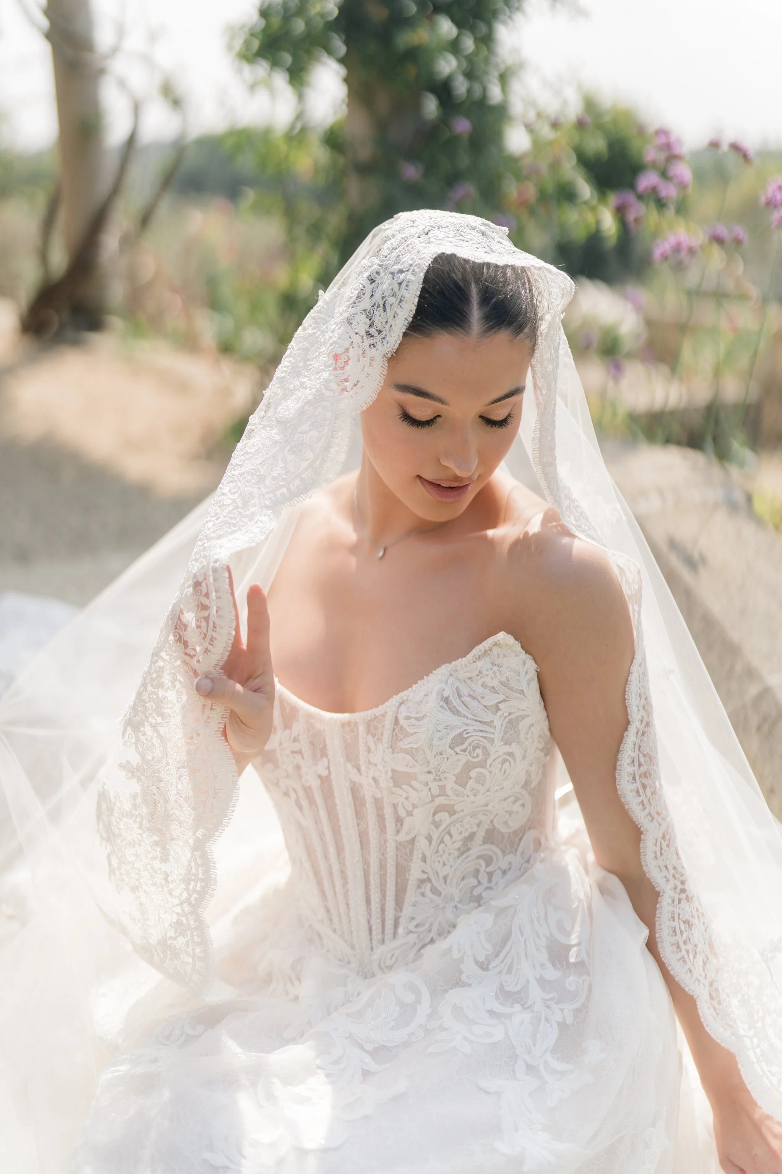 A woman in a white wedding dress with a lace veil outdoors, with trees and purple flowers in the background. Styled shoot held at Euridge Manor assisted by Bootique Weddings and Events.