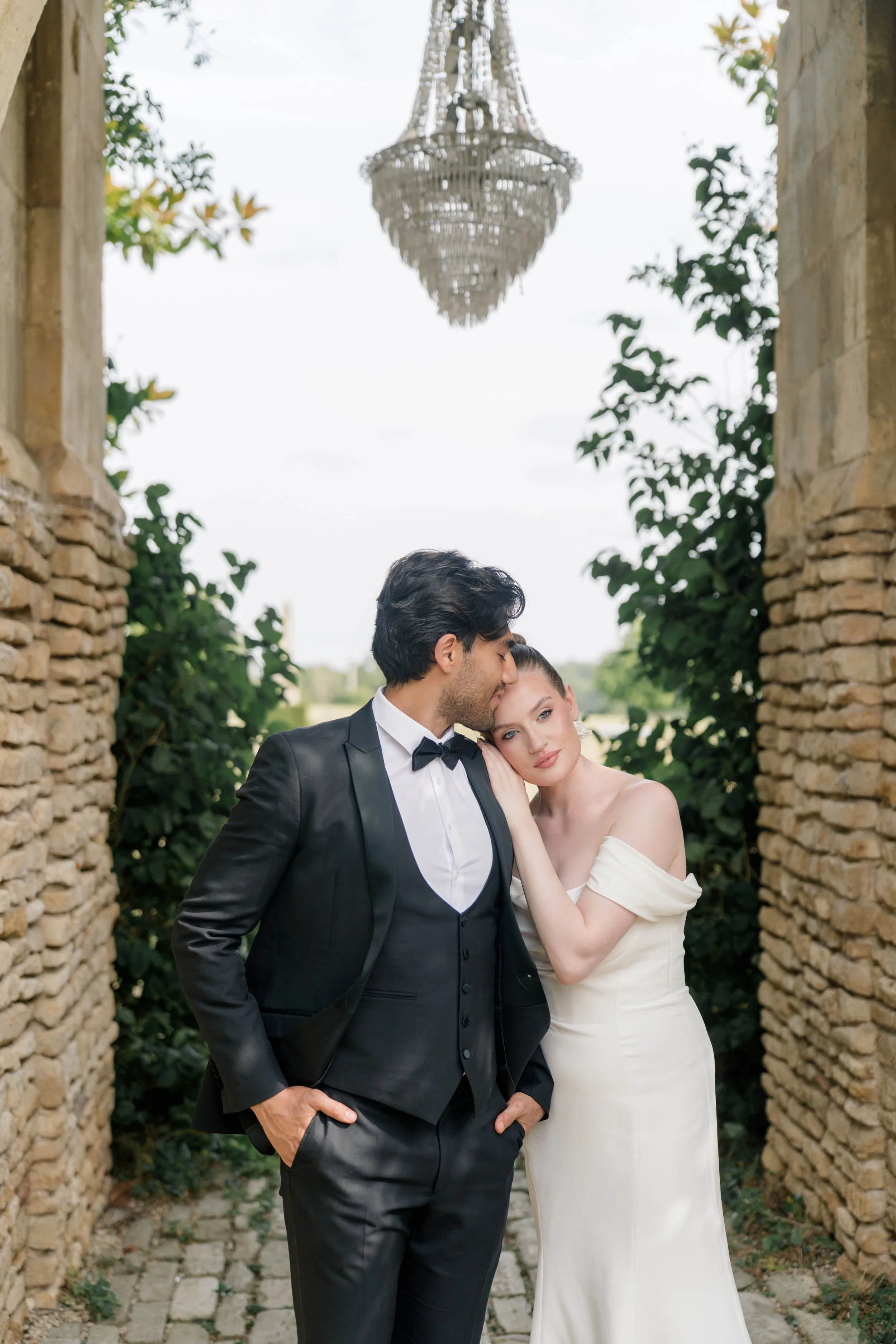 A man in a black tuxedo with a bowtie and a woman in a white off-shoulder gown standing close together outdoors near stone walls and greenery, with a chandelier hanging above. Styled shot held at Euridge Manor assisted by Bootique Weddings and Events