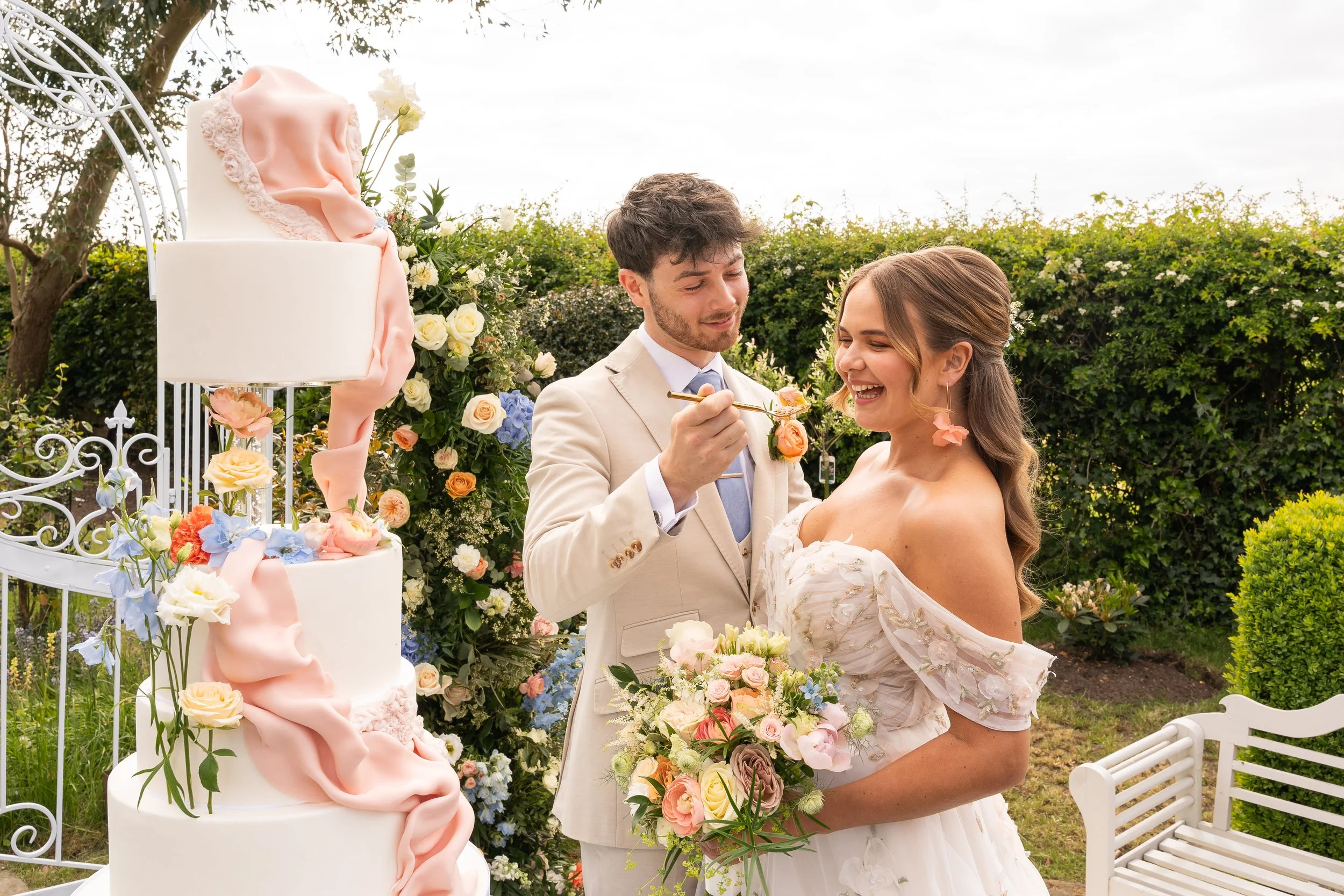 A newlywed couple at their outdoor wedding, with the groom applying blush to the bride's cheek and the bride holding a bouquet of flowers, surrounded by lush greenery and a decorated wedding cake with flowers.