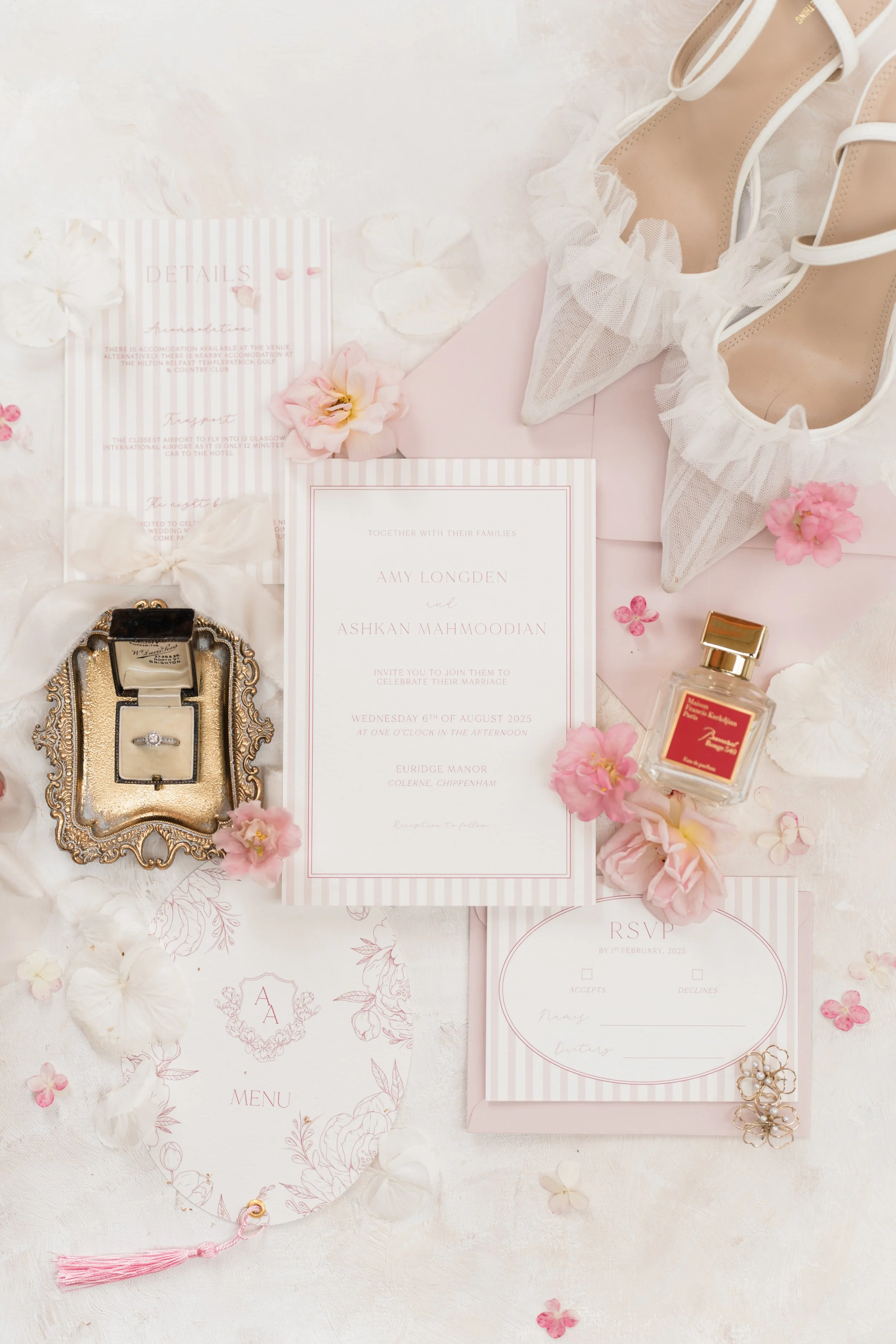 Wedding invitation, jewelry box with engagement ring, pink and white flowers, high heels with ruffled toes, perfume bottle, RSVP card, menu card, and pink envelope arranged on a soft surface for a wedding.