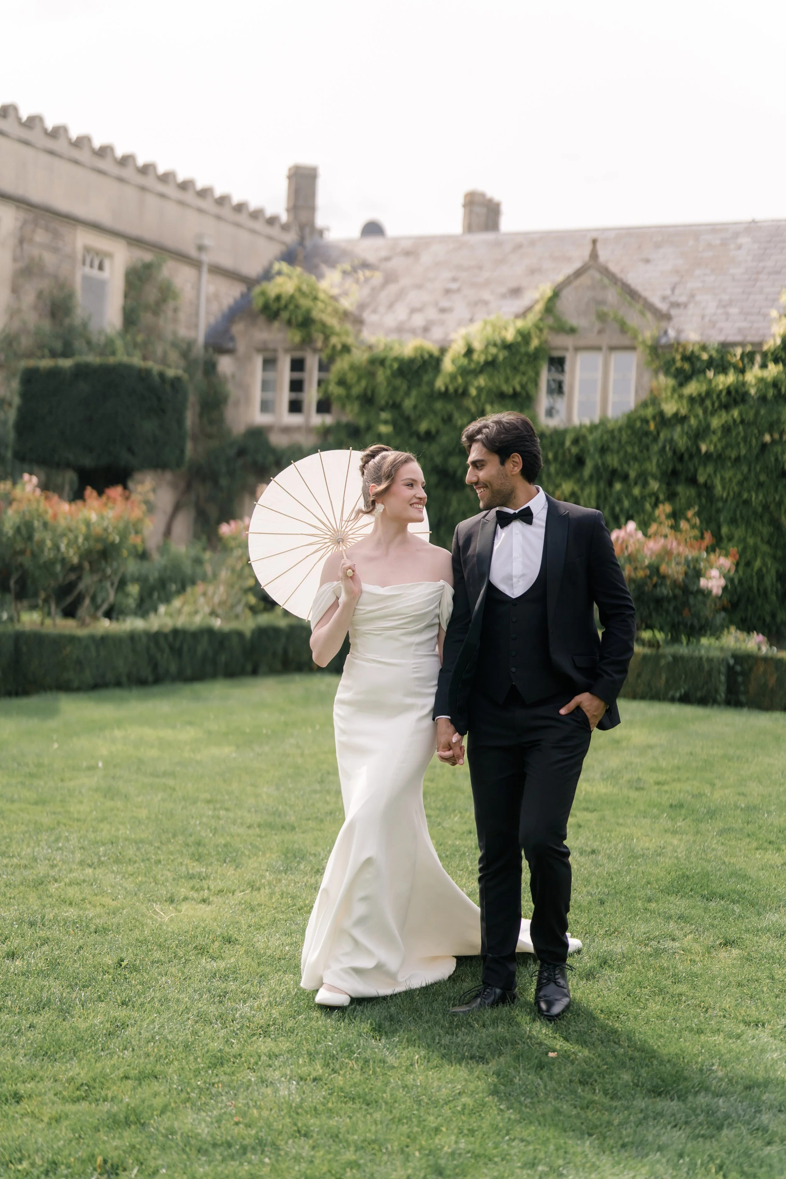 A bride and groom walking hand in hand on a lawn, smiling at each other, with the bride holding a parasol, in front of a historic stone building covered with ivy. Styled shoot held at Euridge Manor assisted by Bootique Weddings and Events.