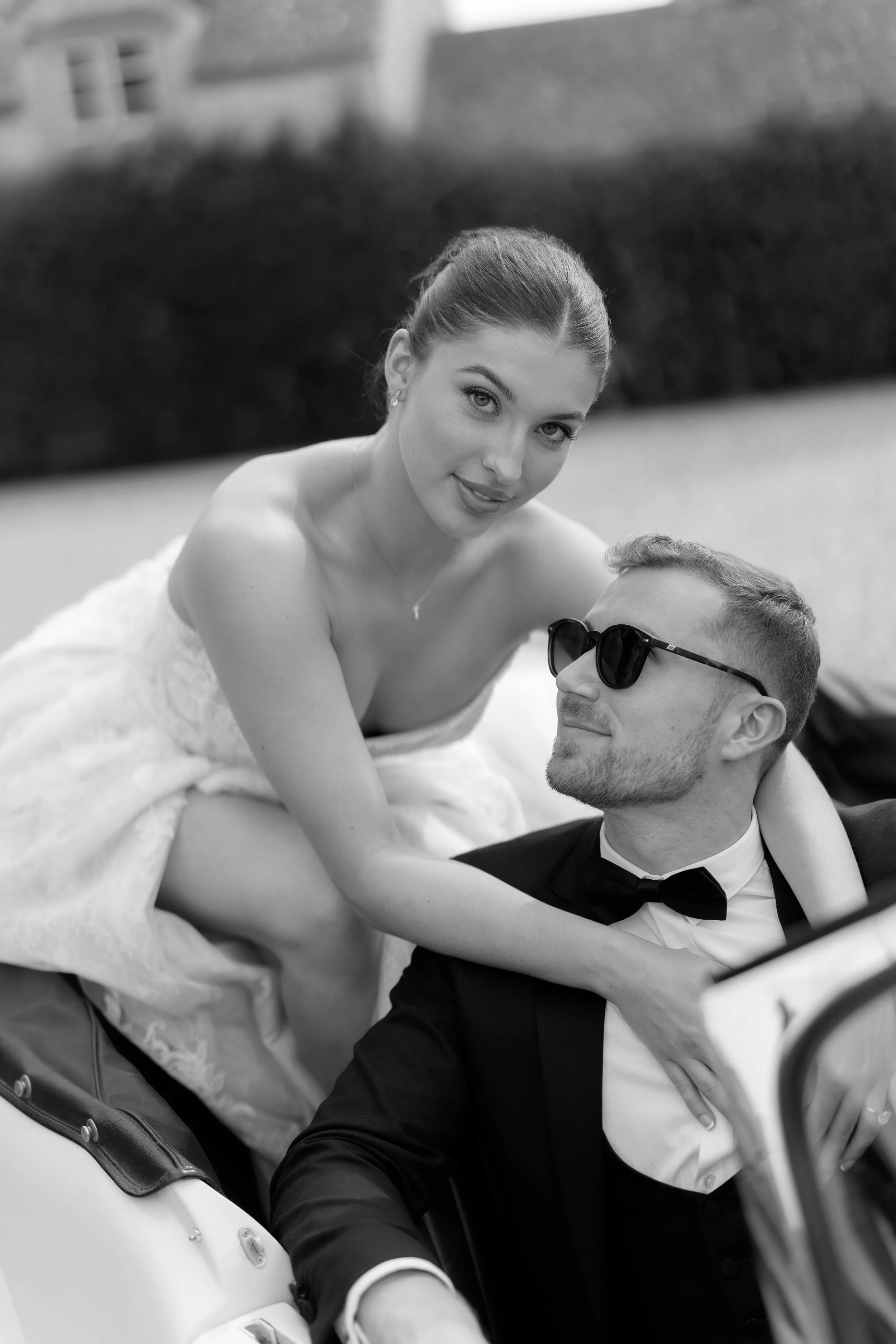 Black and white photo of a woman in a wedding dress and a man in a tuxedo with sunglasses sitting in a vintage car, outdoors. Styled shoot held at Euridge Manor assisted by Bootique Weddings and Events.