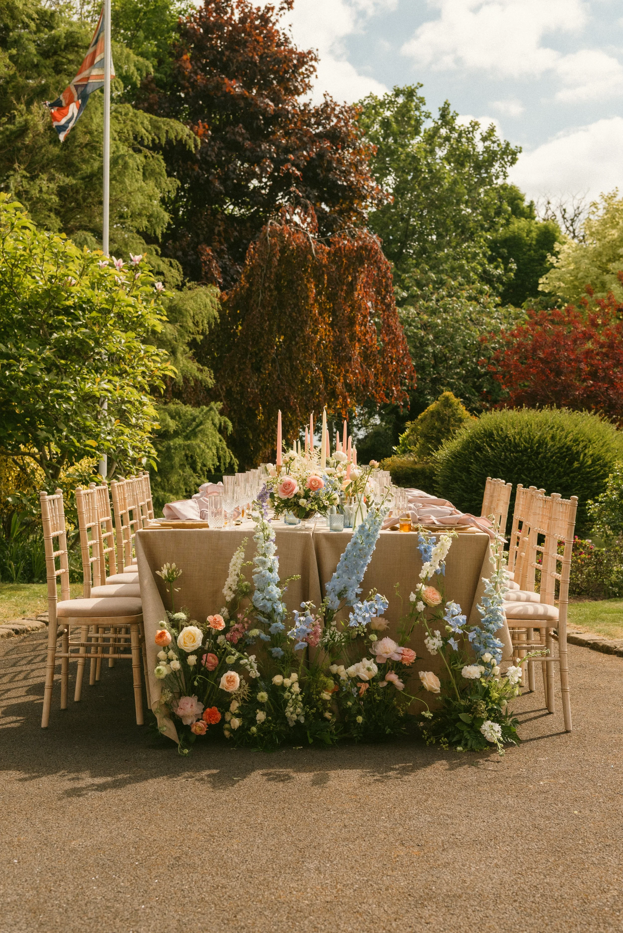 An outdoor dining table set for a celebration with floral arrangements, candles, and dinnerware, surrounded by lush greenery. Taken at a styled shoot at The Cheshire Venue and planned by Bootique Weddings and Events.
