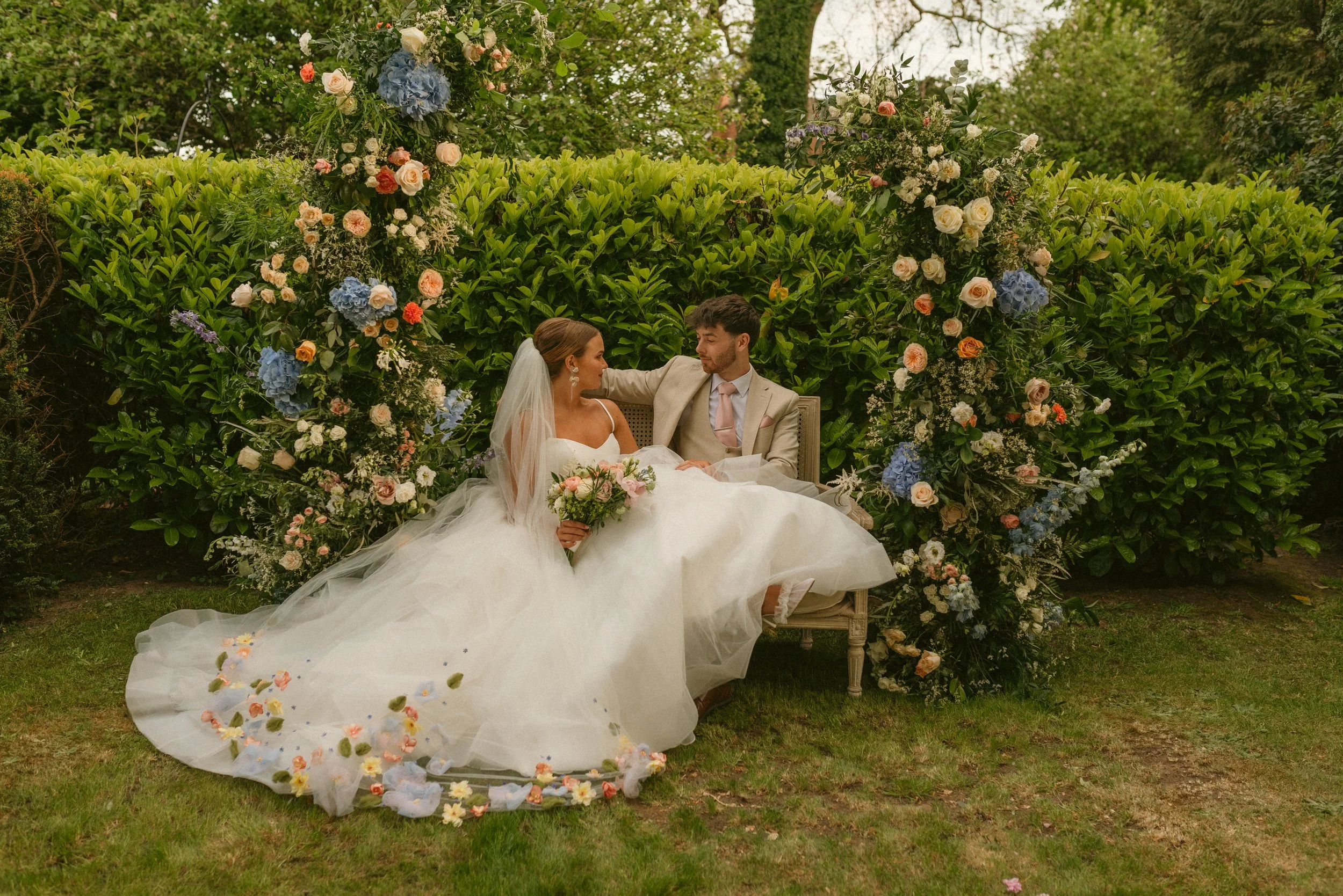 A bride and groom sitting on a bench in front of a floral arch at an outdoor wedding. The bride is in a white wedding gown holding a bouquet, and the groom is in a suit with a pink tie, gazing at each other.