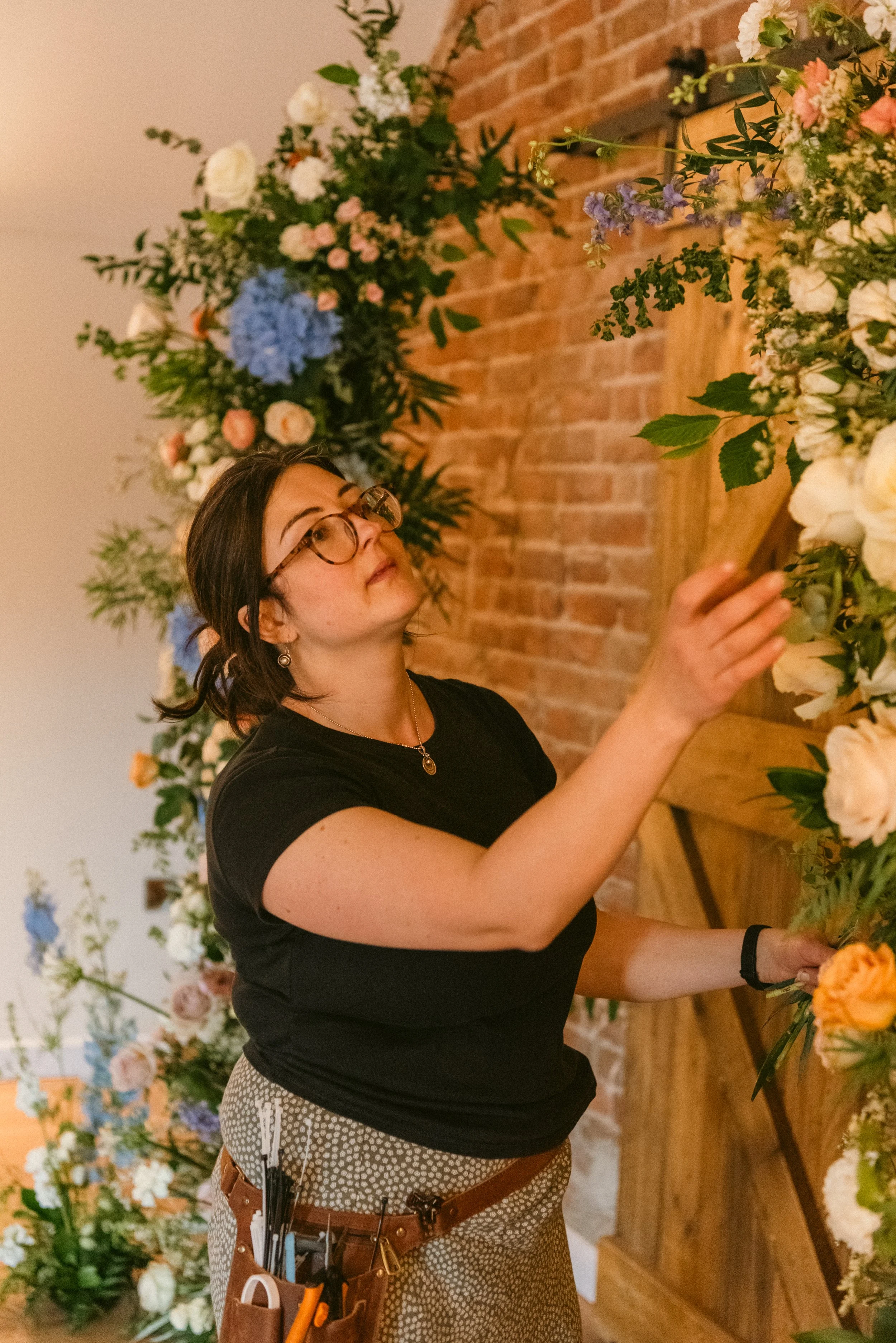 Woman arranging flowers in a floral display, with a brick wall background. Taken at a styled shoot at The Cheshire Venue and planned by Bootique Weddings and Events.