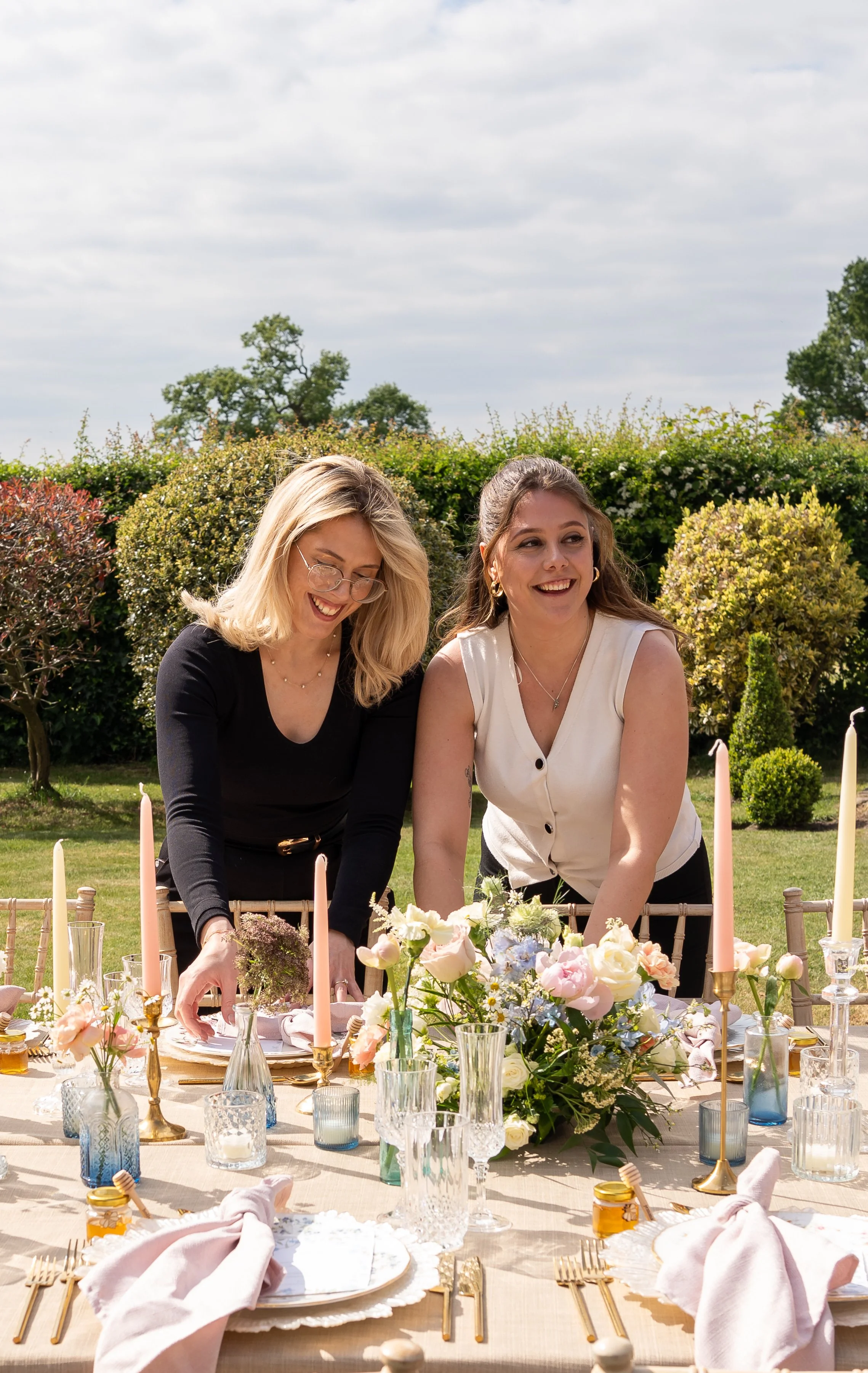 Two women are setting a table outdoors with flowers, candles, and glassware, smiling and laughing in a garden. Styled shoot held at The Cheshire Venue planned by wedding planner Bootique Weddings and Events.