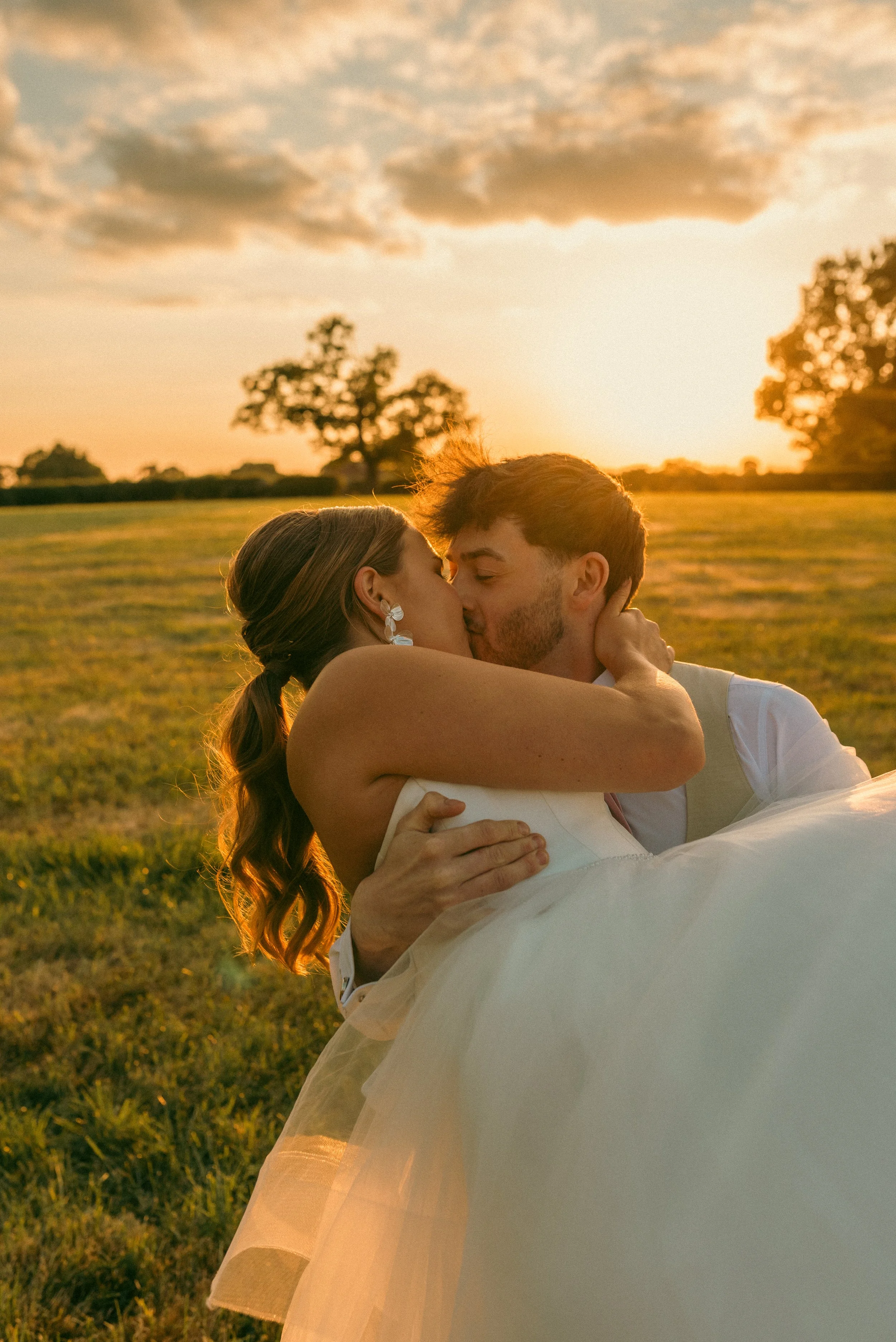 A couple in wedding attire kissing outdoors during sunset, with a grassy field and trees in the background. Styled shoot held at The Cheshire Venue planned by wedding planner Bootique Weddings and Events.