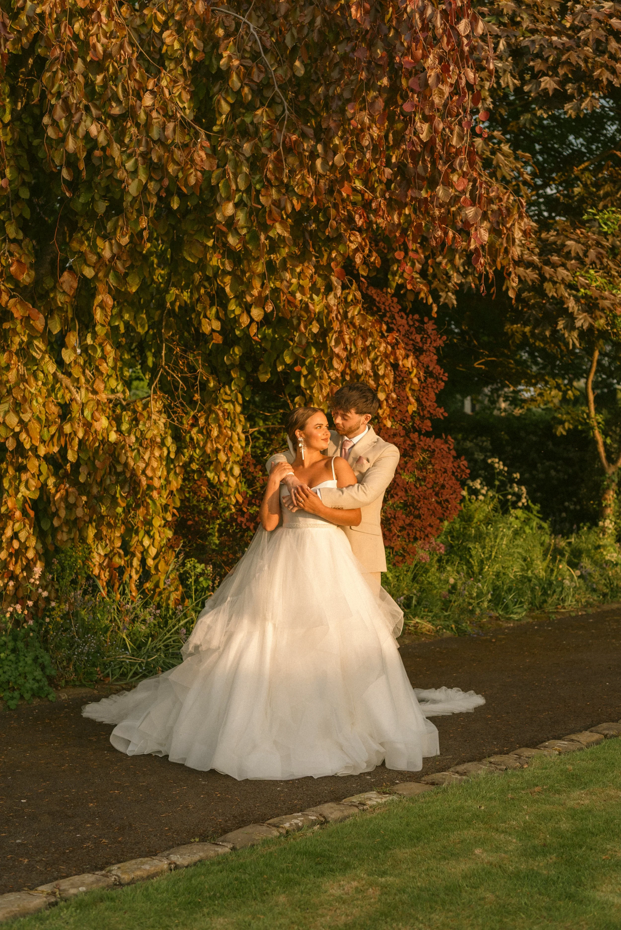 A bride in a white wedding gown and a groom in a beige suit standing close together in a garden with autumn-colored foliage, embracing each other lovingly. Styled shoot held at The Cheshire Venue planned by wedding planner Bootique Weddings and Event