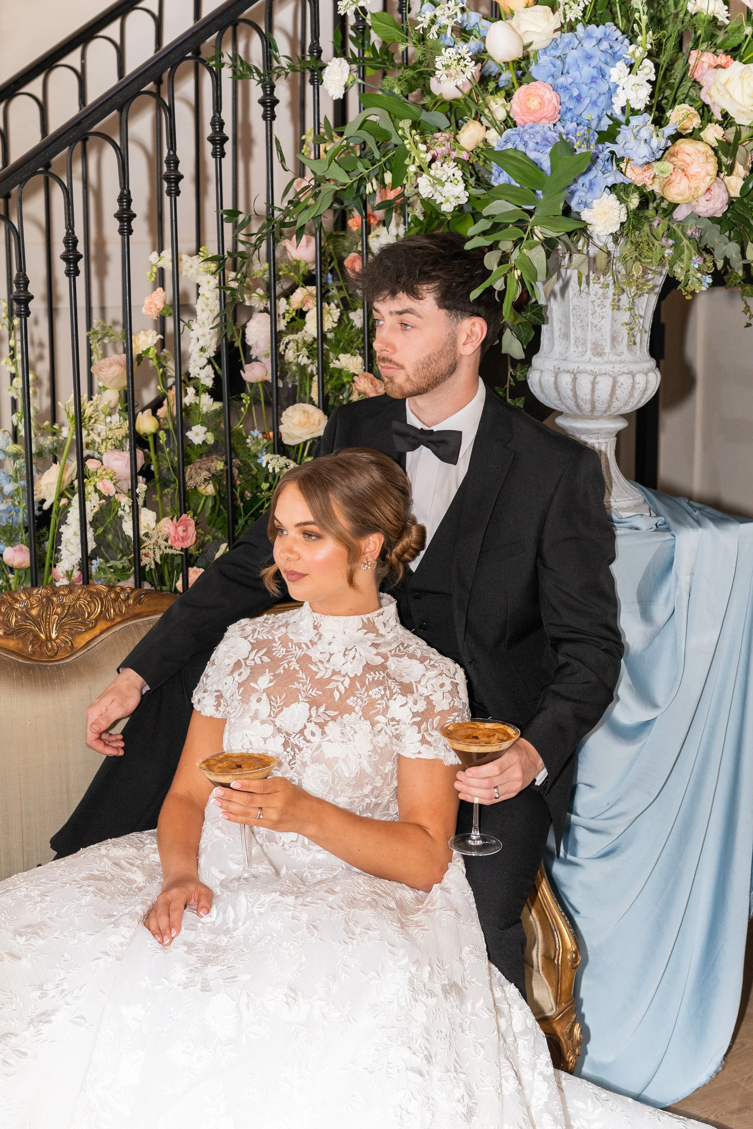 A bride and groom at their wedding reception, seated on an ornate sofa with the bride holding a drink and the groom standing behind her, surrounded by a large floral arrangement.