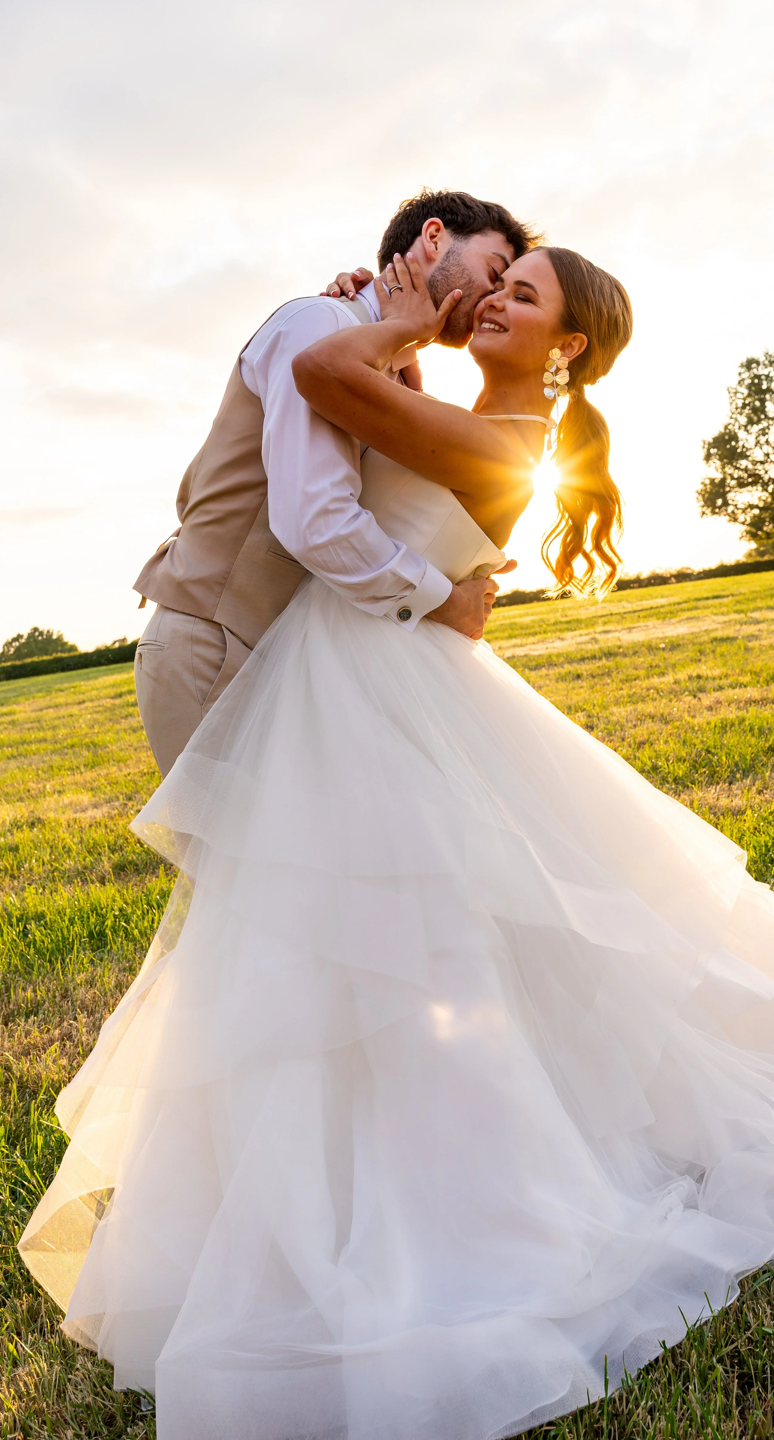 A happy couple in wedding attire sharing a joyful embrace outdoors at sunset, with the bride wearing a white gown and the groom in a beige vest and pants. Styled shoot held at The Cheshire Venue planned by wedding planner Bootique Weddings and Events