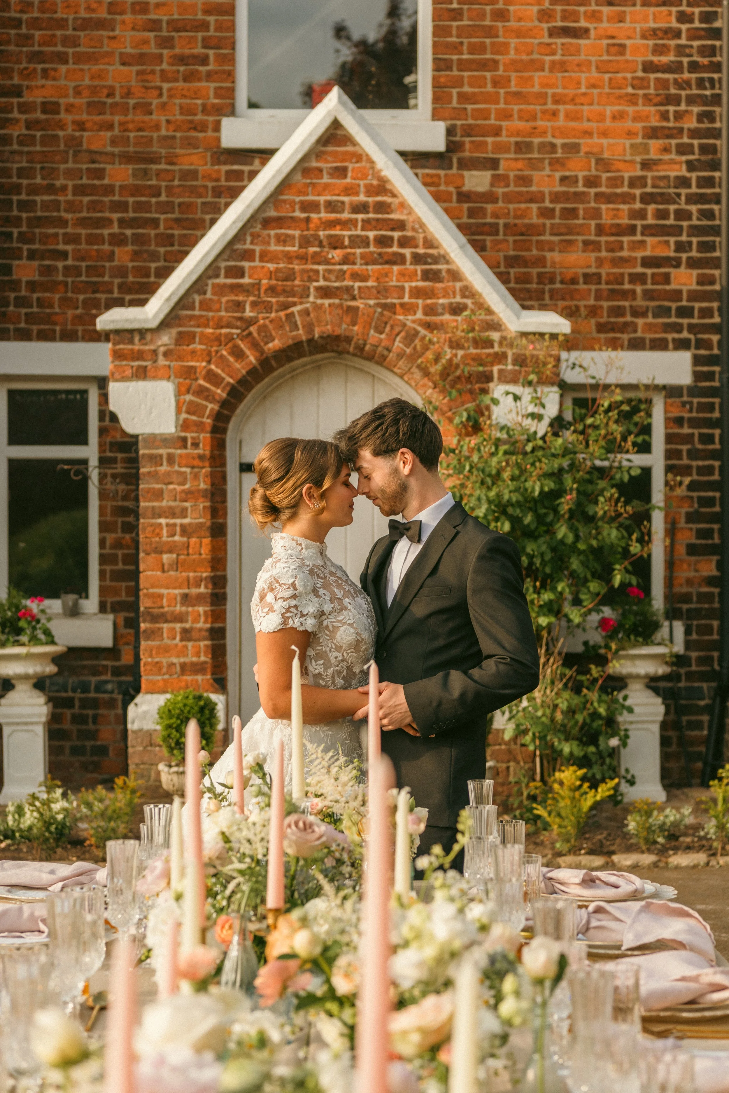 A bride and groom stand close together with foreheads touching at their wedding reception outside a brick building, surrounded by flowers and candles on the table. Taken at a styled shoot at The Cheshire Venue and planned by Bootique Weddings.