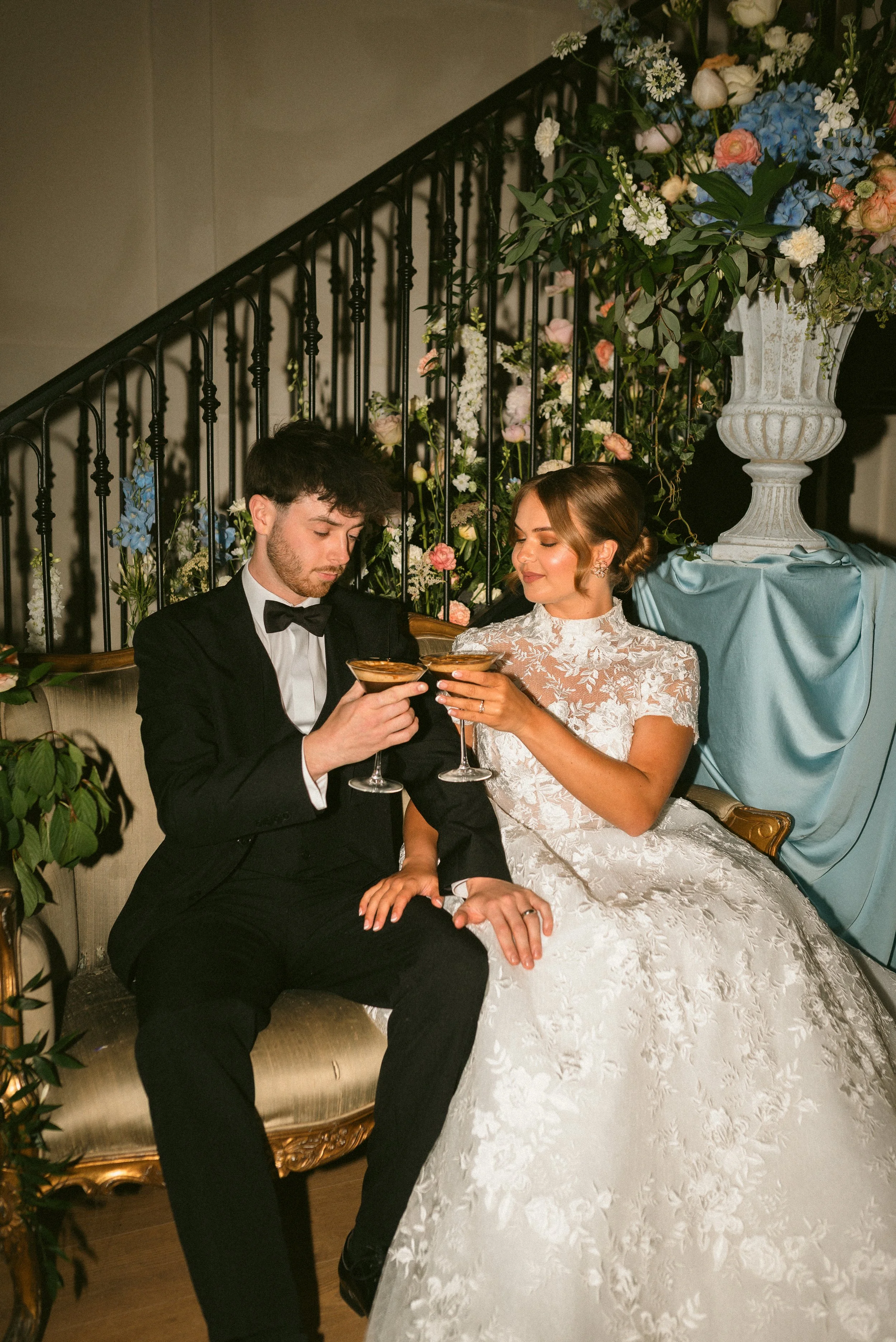 A bride and groom in formal attire sitting on a vintage sofa, holding glasses of champagne, and toasting each other at their wedding reception, with floral decorations in the background. Styled shoot held at The Cheshire Venue.
