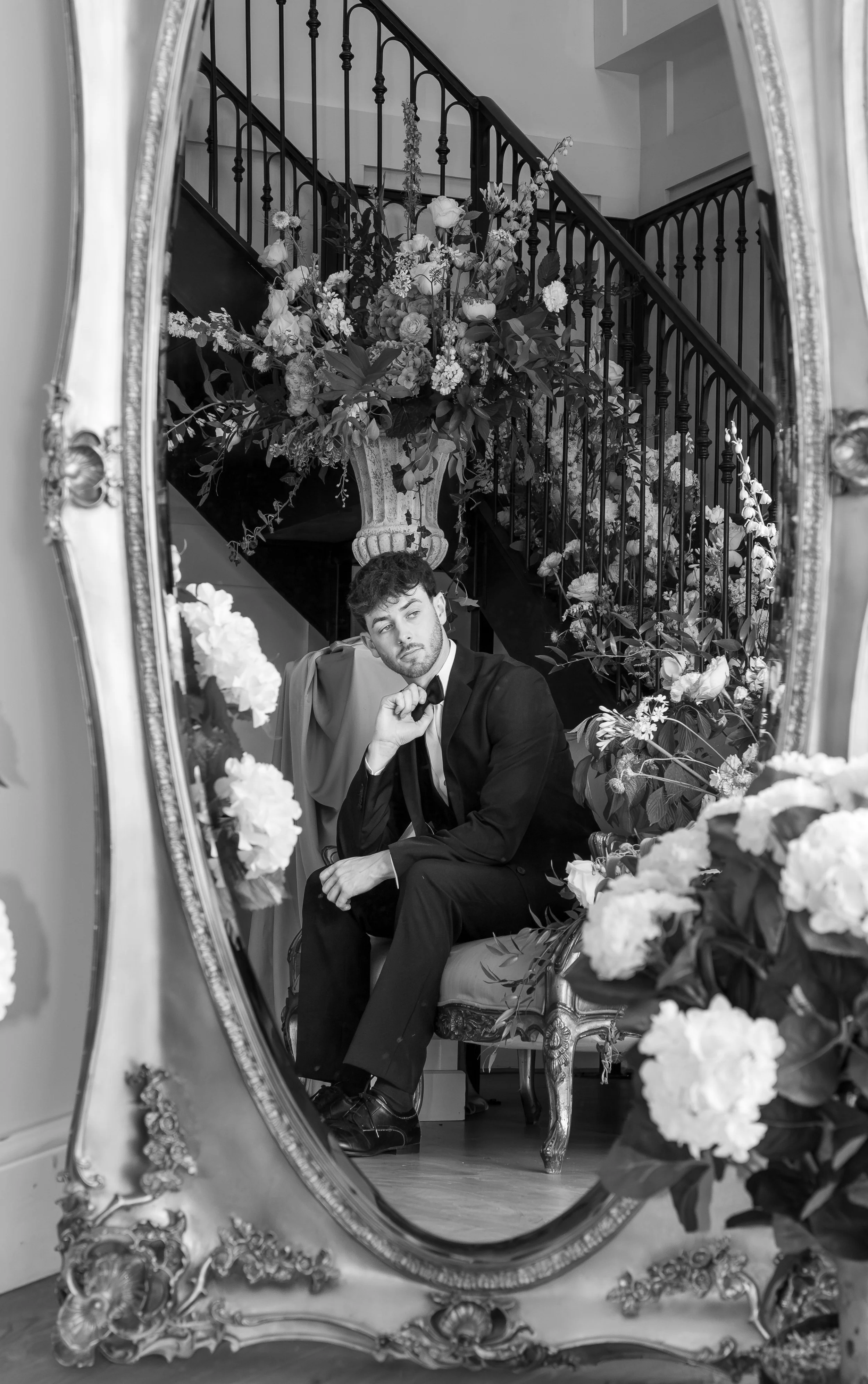 A young man in a tuxedo taking a selfie in an ornate mirror decorated with flowers, with a staircase and flowers in the background. Styled shoot held at The Cheshire Venue planned by wedding planner Bootique Weddings and Events.
