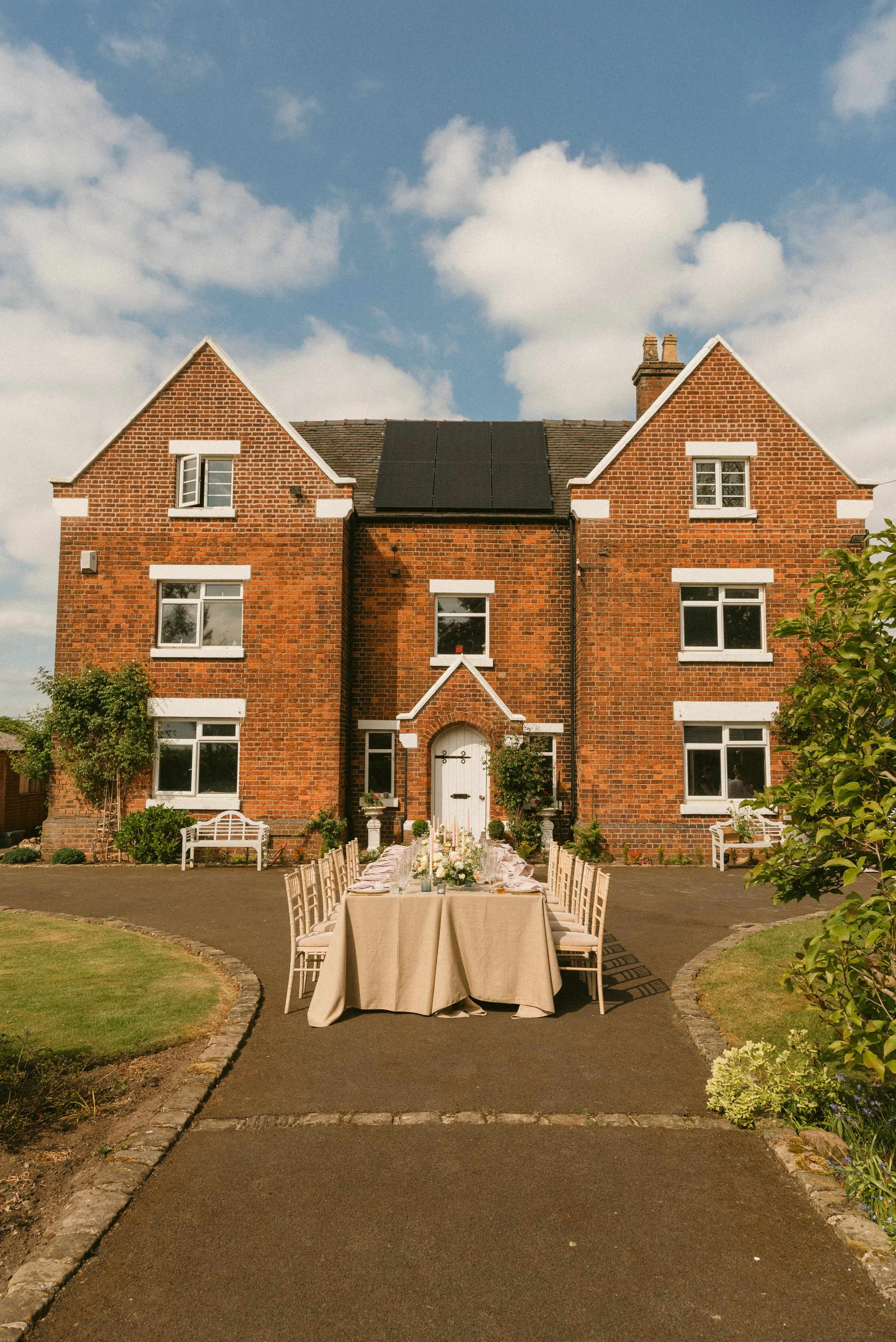 Outdoor party setup with a long table and chairs in front of a large brick house with multiple windows, on a sunny day with a partly cloudy sky. Taken at a styled shoot at The Cheshire Venue and planned by Bootique Weddings and Events.