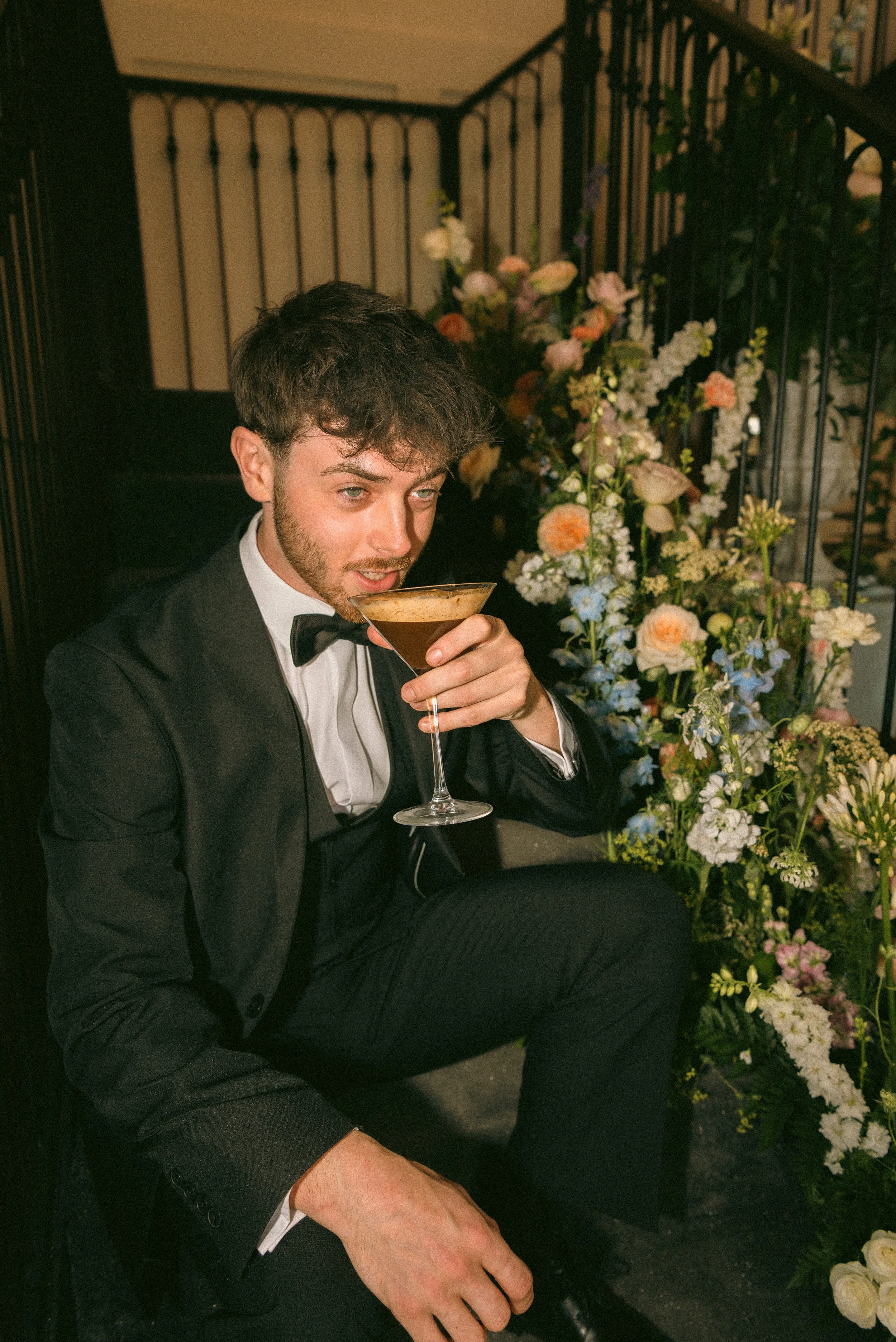 A man in a tuxedo drinking a cocktail at an indoor event with floral arrangements in the background. Styled shoot held at The Cheshire Venue planned by wedding planner Bootique Weddings and Events.
