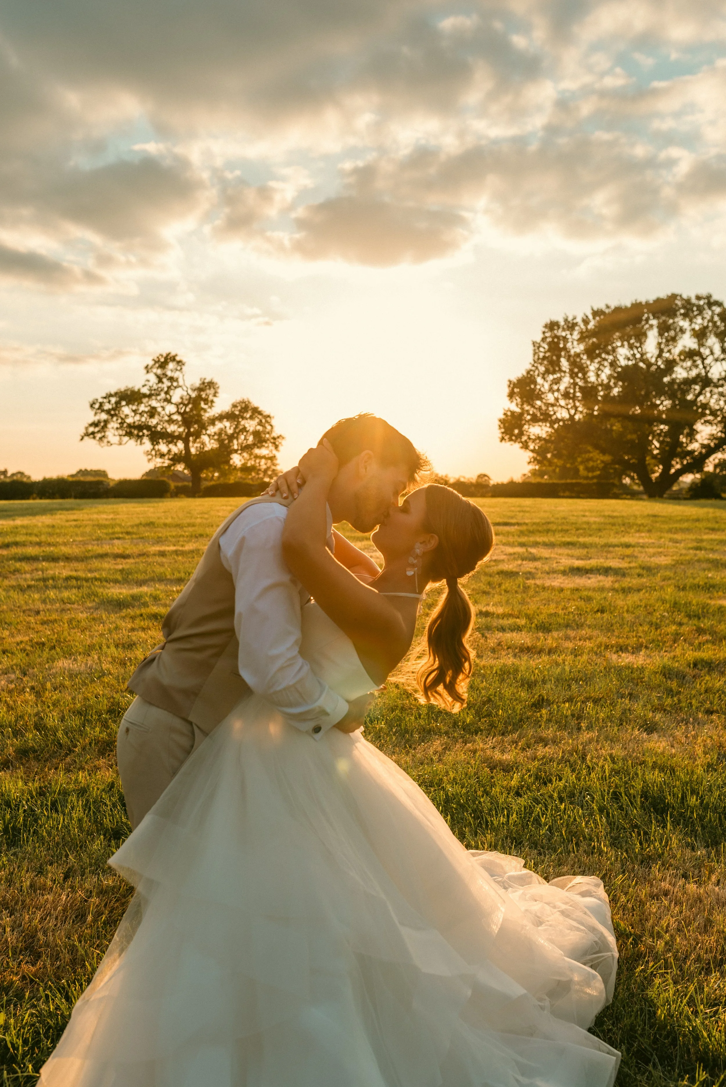 A bride and groom sharing a kiss in a sunlit field at sunset, with trees in the background. Styled shoot held at The Cheshire Venue planned by wedding planner Bootique Weddings and Events.