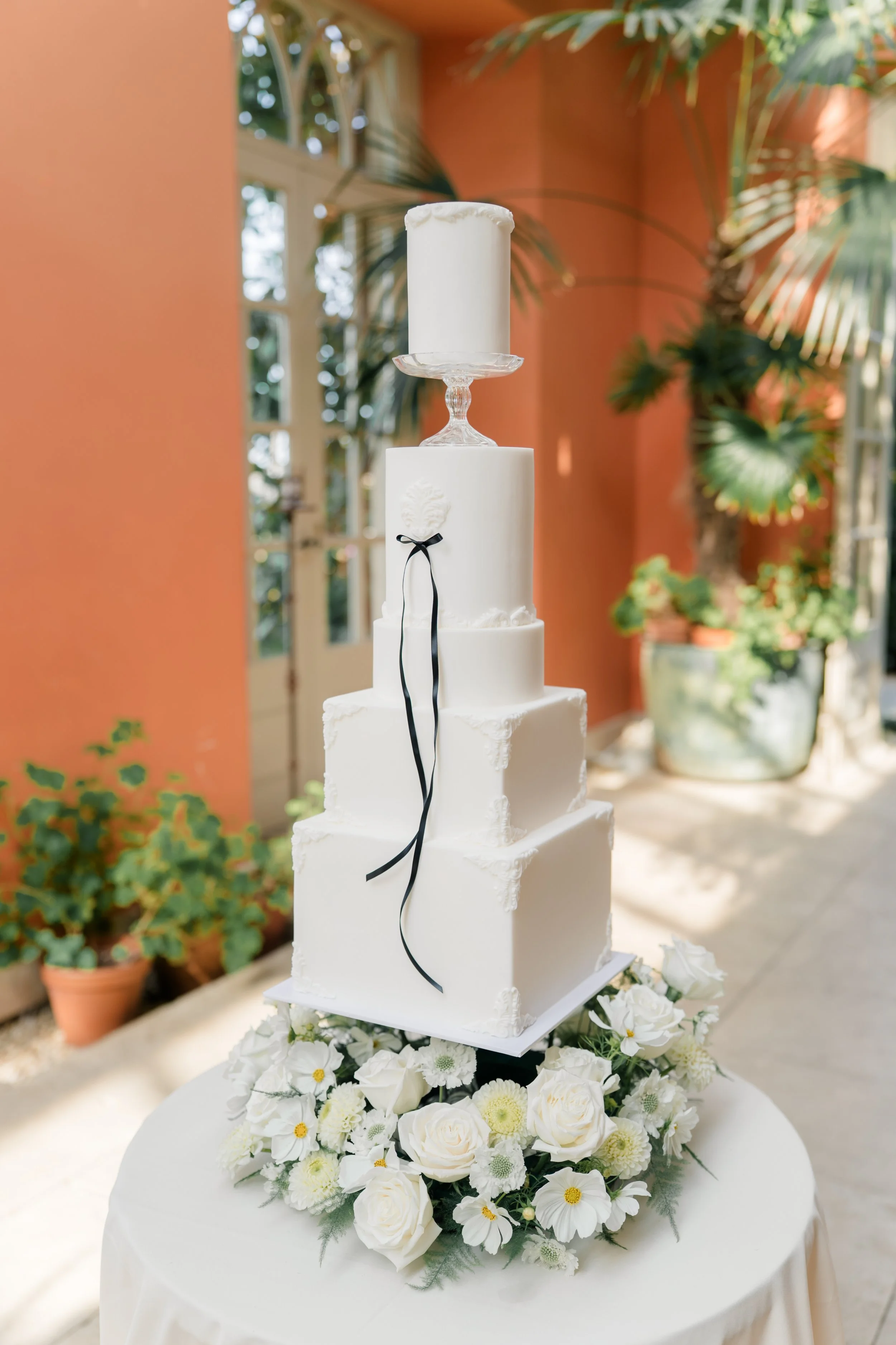 A multi-tiered white wedding cake with a white candle on top, decorated with a black ribbon and floral details, surrounded by white flowers at its base on a table with a white tablecloth indoors with plants and windows in the background.