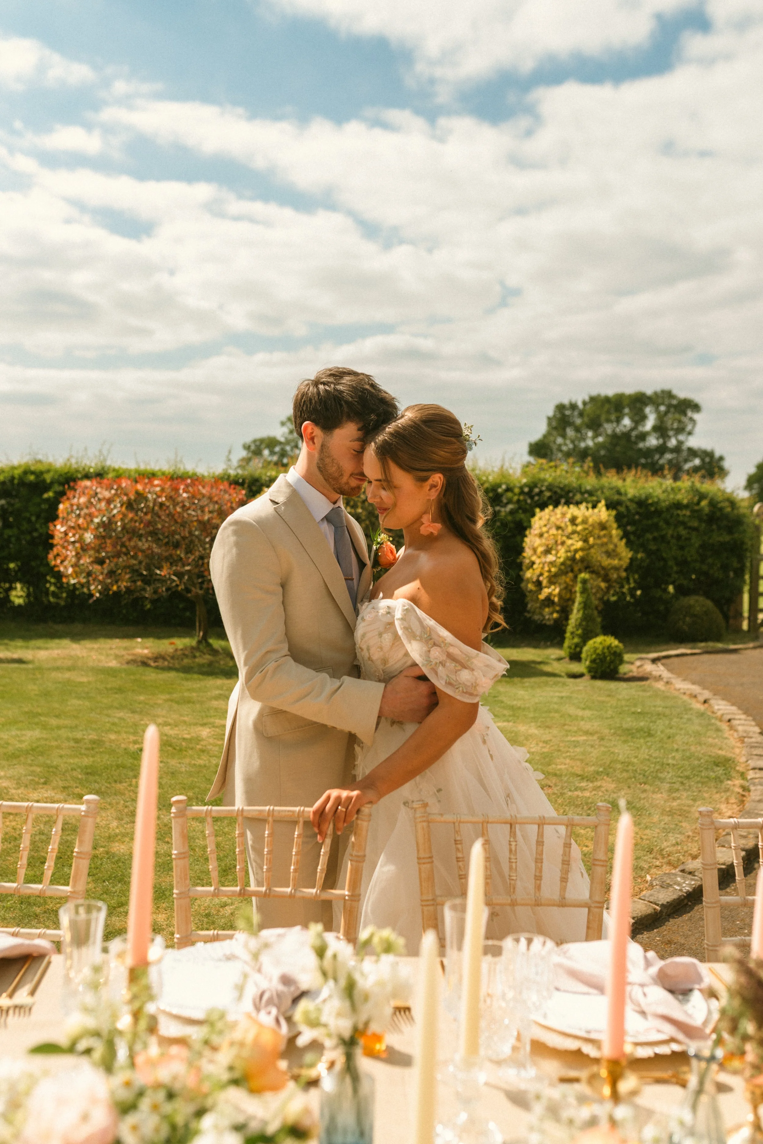 A bride and groom embrace at an outdoor wedding reception, standing behind a decorated table with flowers and candles, under a partly cloudy sky with lush greenery in the background. Taken at a styled shoot planned by Bootique Weddings and Events.