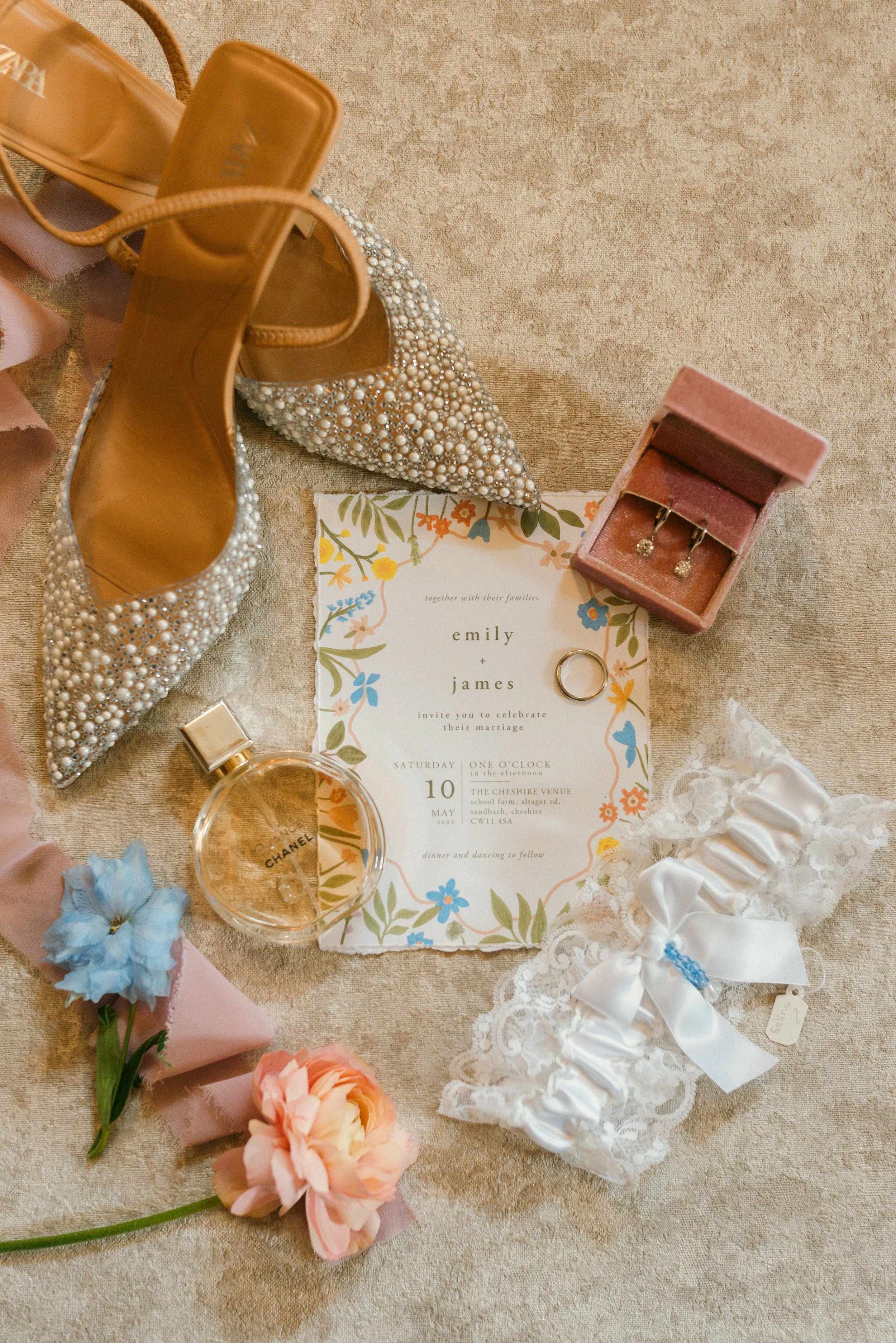 Wedding invitation surrounded by bridal accessories including pearl-embellished high heels, lace garter with bow, jewelry box with earrings, a perfume bottle, wedding rings, and colorful flowers on a beige surface.