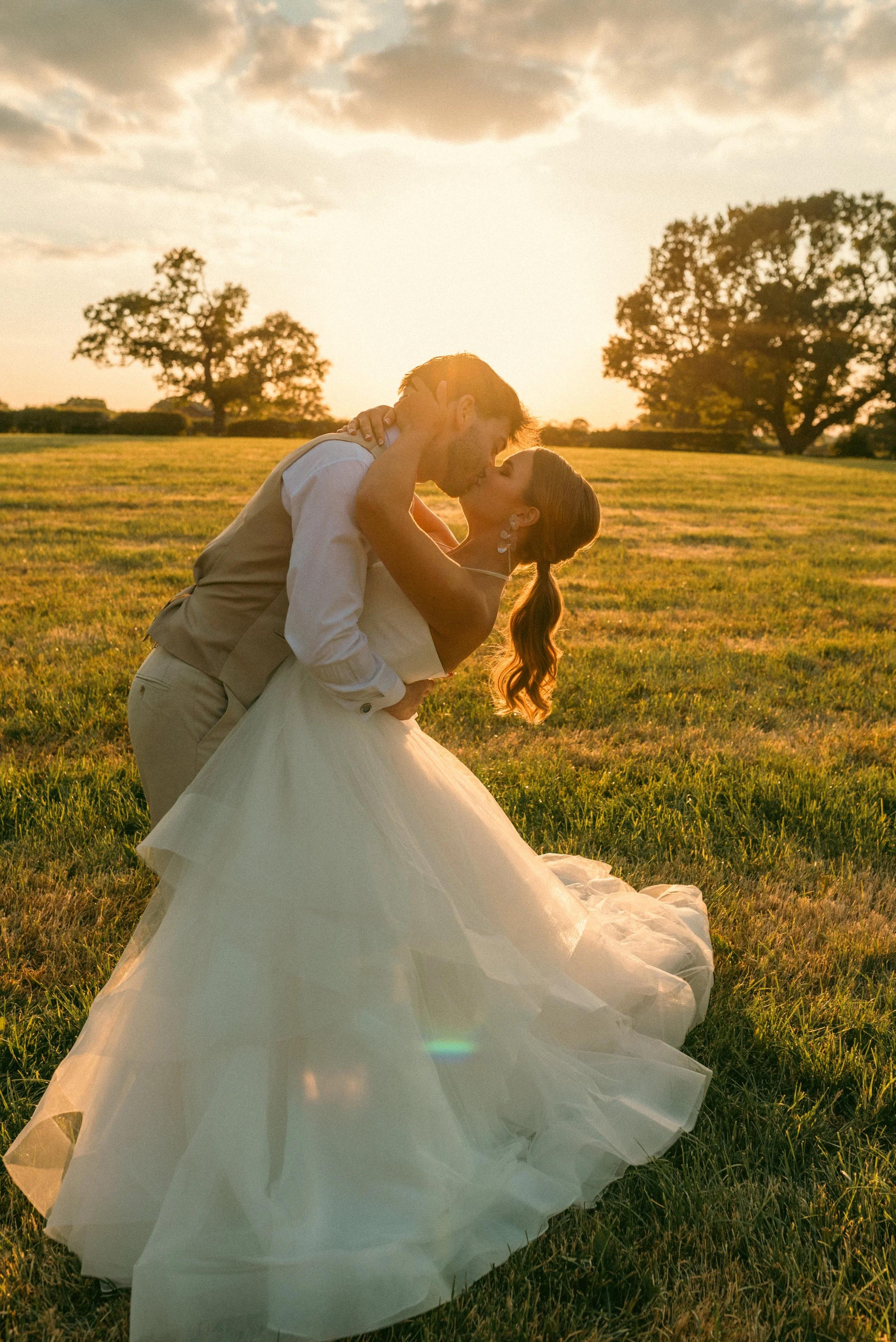 A newlywed couple sharing a kiss in a field during sunset, with large trees in the background and the bride wearing a white wedding gown. Styled shoot held at The Cheshire Venue planned by wedding planner Bootique Weddings and Events.