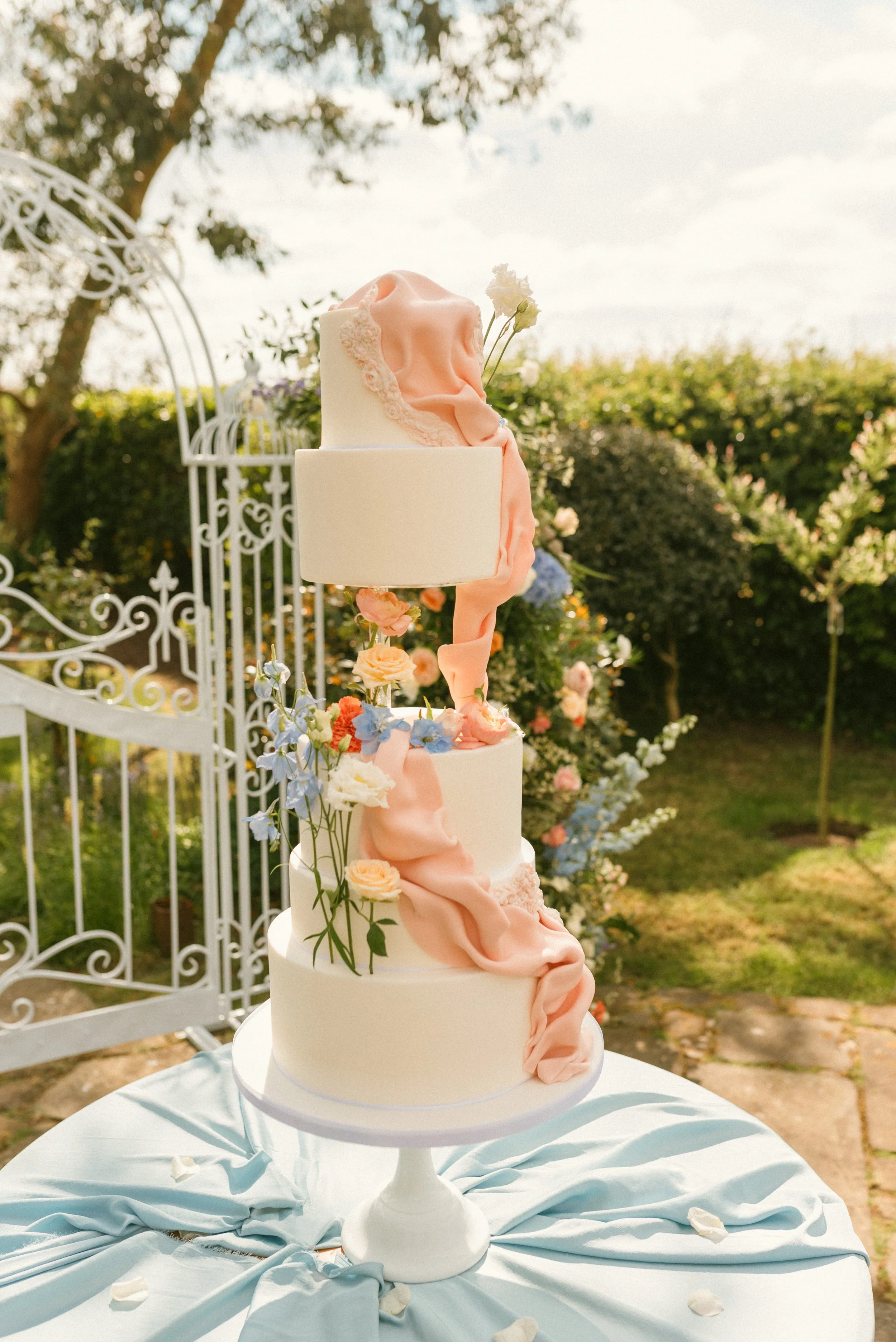 A multi-tier wedding cake decorated with pink and white fabric-like fondant and fresh flowers, set outdoors on a white stand with a draped light blue cloth and flower petals, with a garden background.