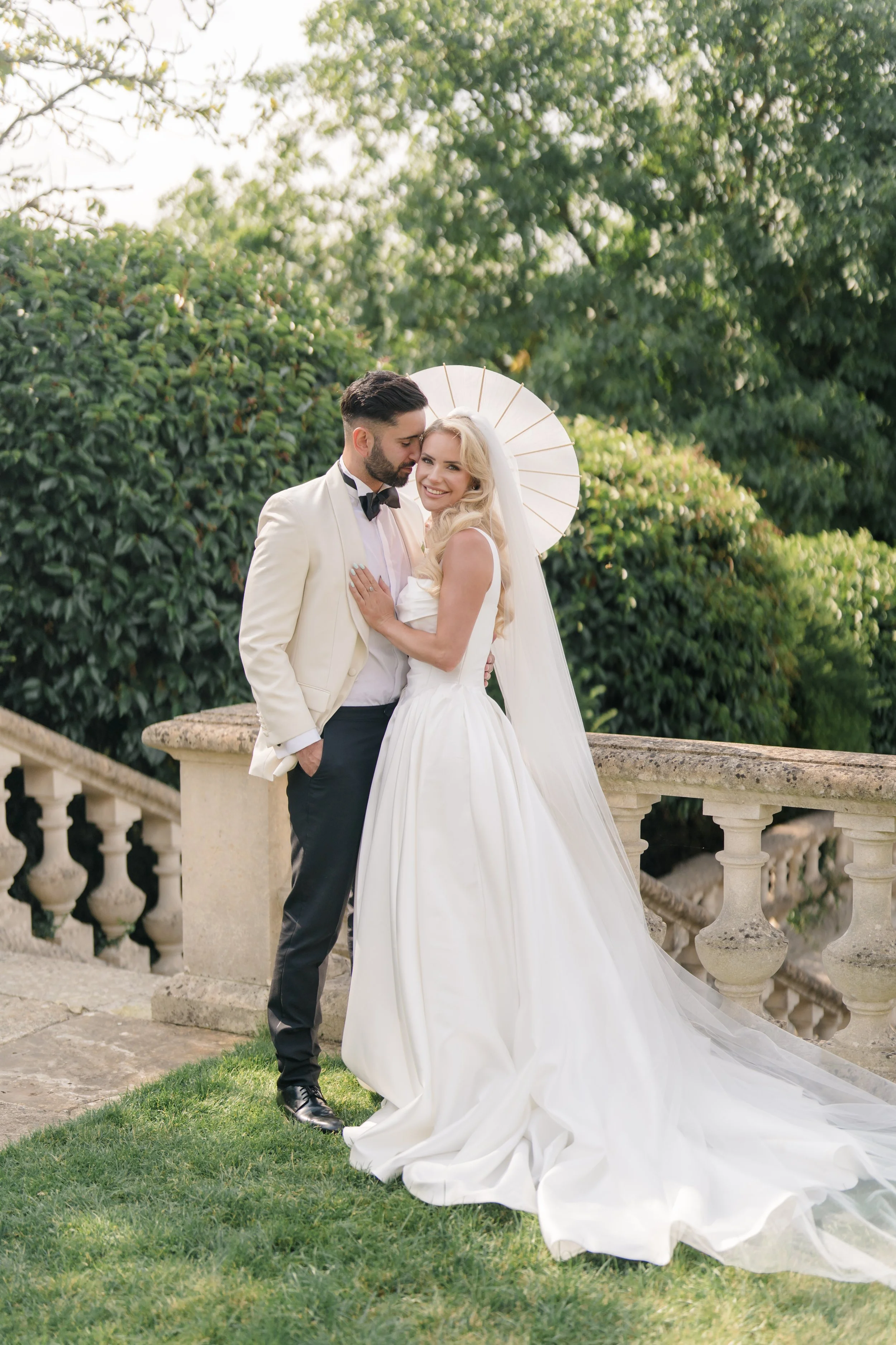A smiling bride and groom in wedding attire pose outdoors next to a stone railing, with lush green trees in the background. The bride is wearing a white wedding dress with a long train and holding a white parasol, while the groom is dressed in a tuxe