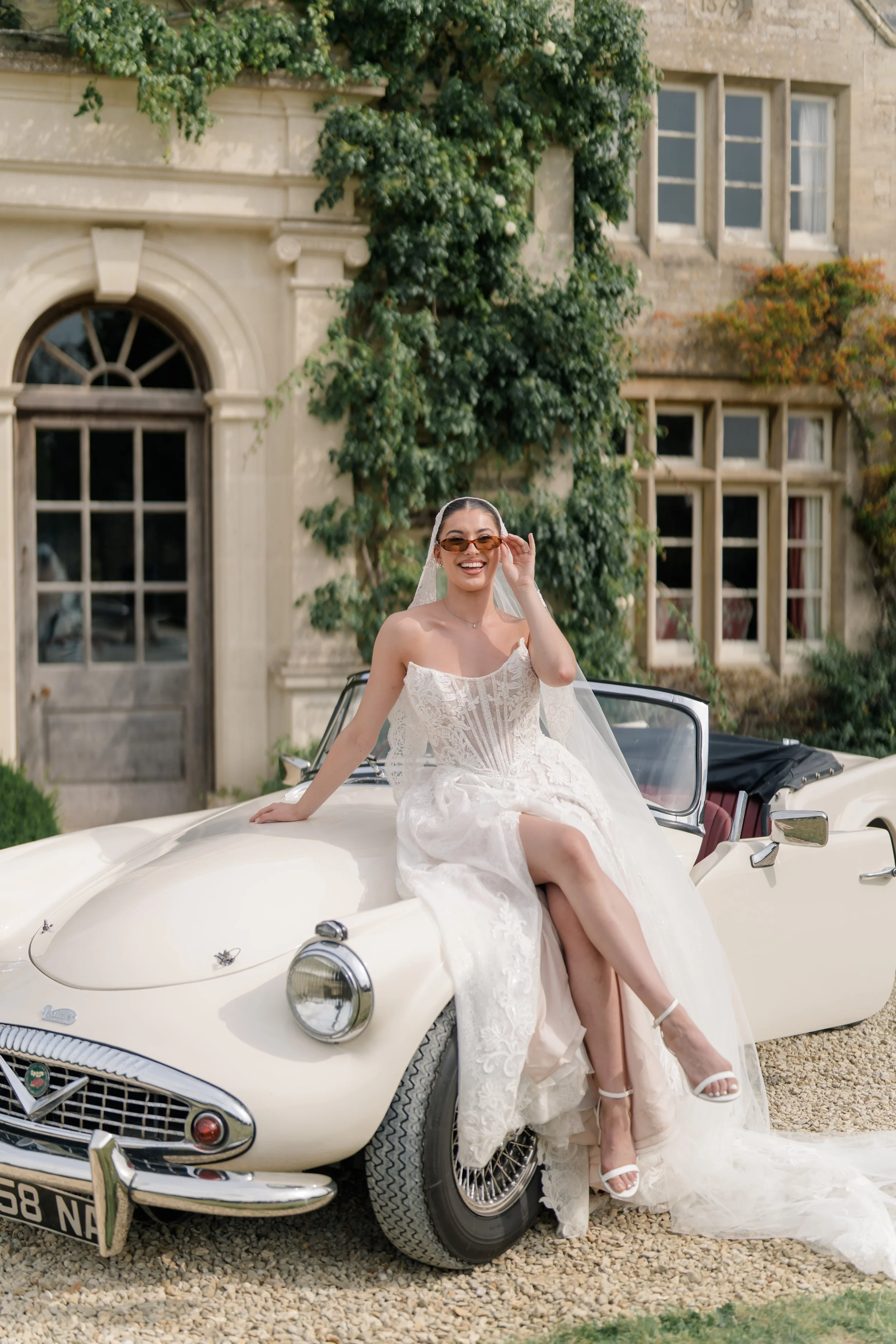 A bride in a white wedding dress sitting on a vintage white convertible car in front of a historic stone building with arched windows and surrounding green foliage, smiling and adjusting sunglasses.