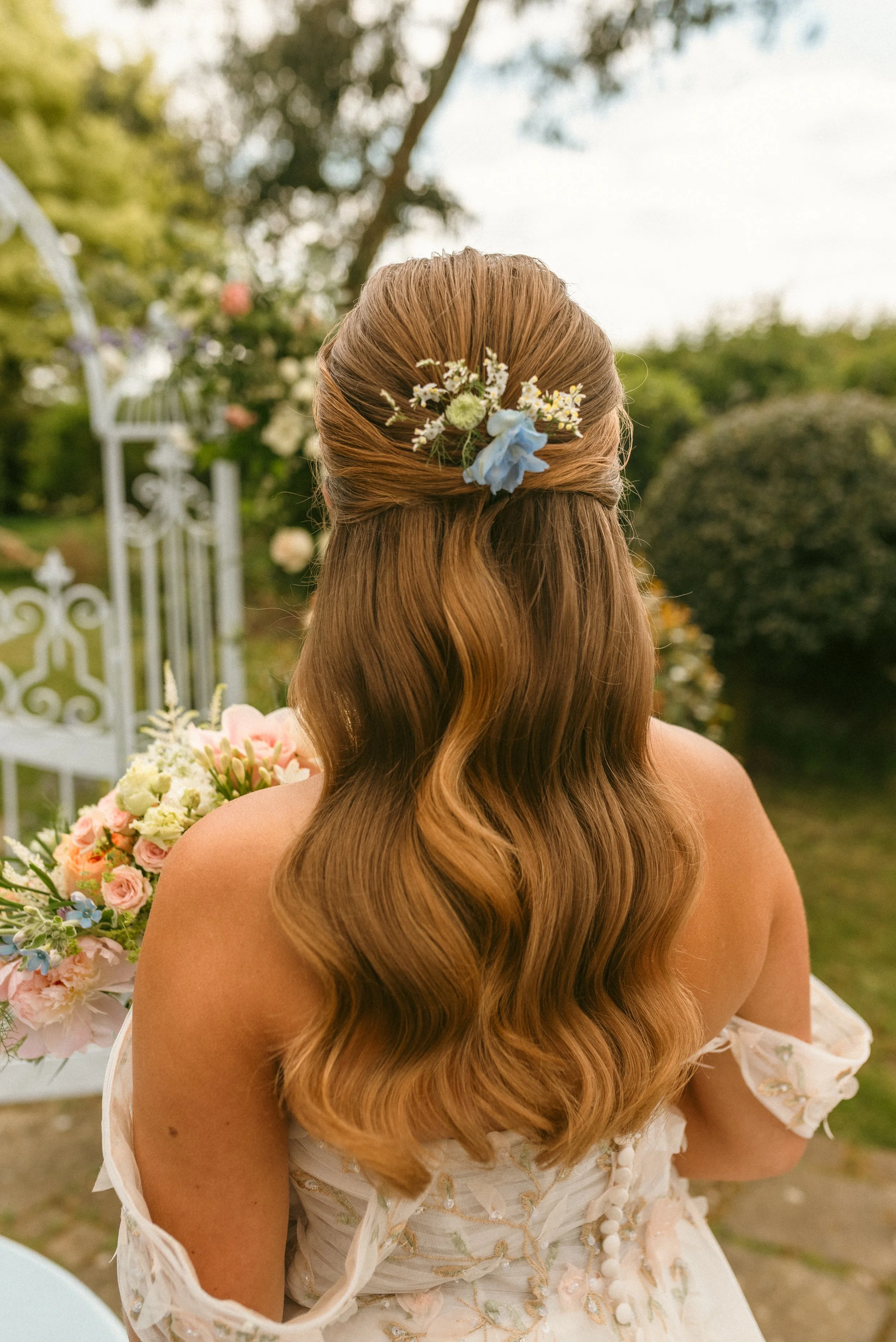 Back view of a woman with long, wavy, brown hair, wearing a white dress with floral embroidery, holding a bouquet of pink, white, and blue flowers, standing outdoors near a garden gate. Taken at a styled shoot planned by Bootique Weddings and Events.
