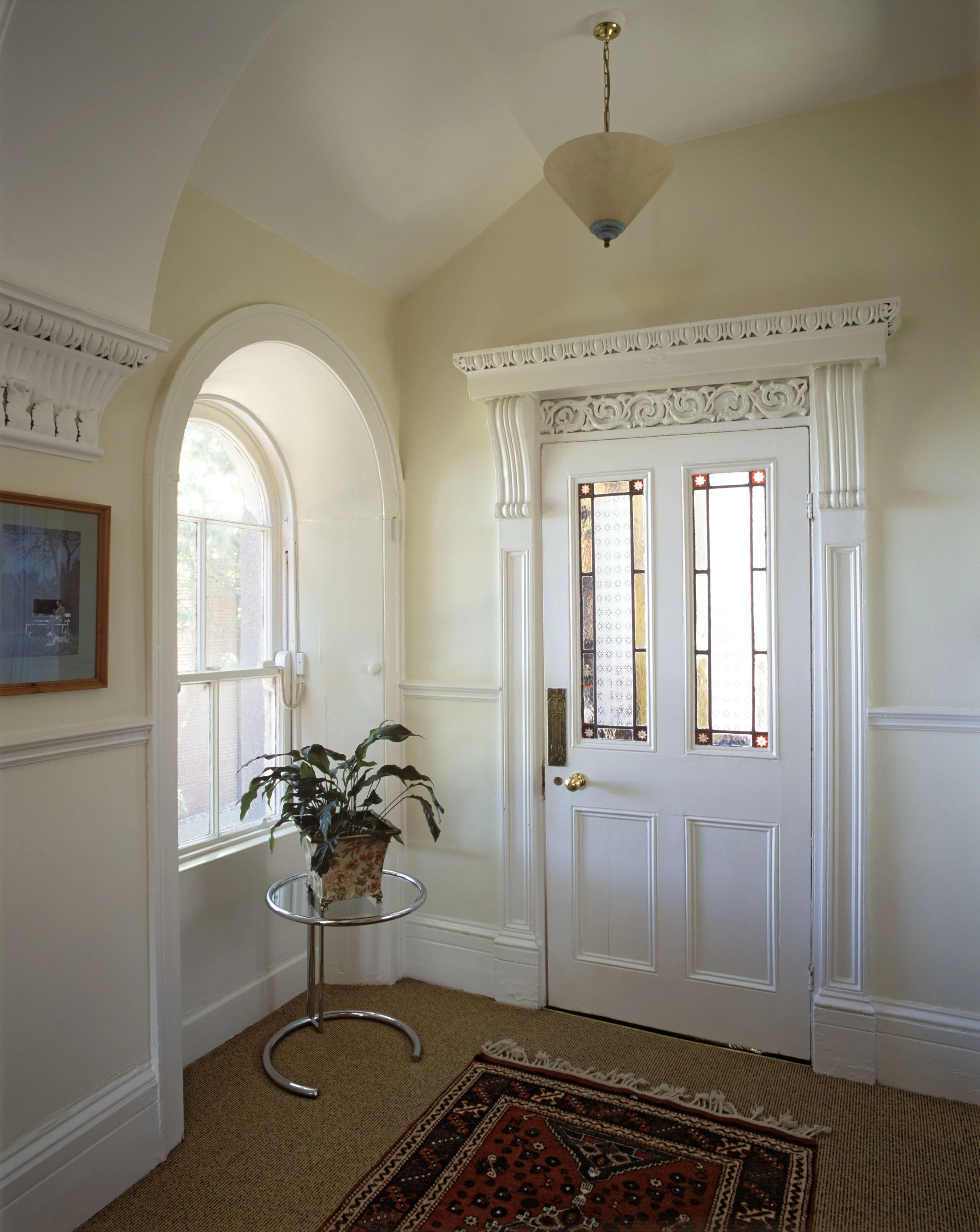 Entryway with ornate white trim, stained glass window on door, potted plant on side table, window with phone, area rug on carpeted floor, ceiling light fixture.