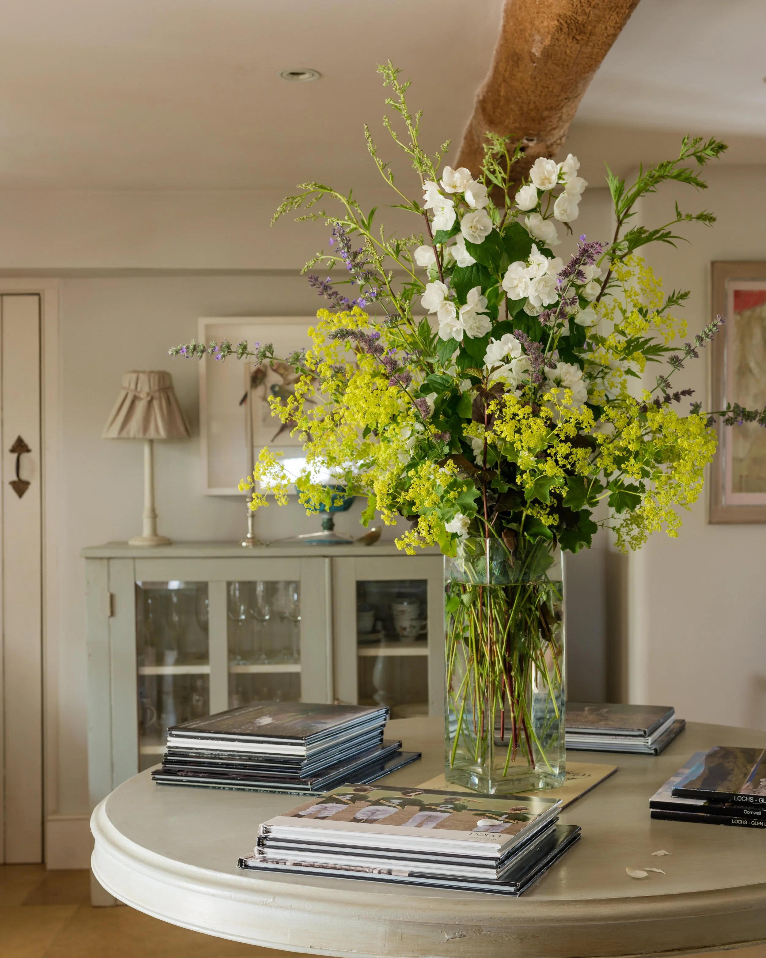 A large glass vase filled with white, yellow, and purple flowers on a round white table with stacks of magazines in a cozy living room.