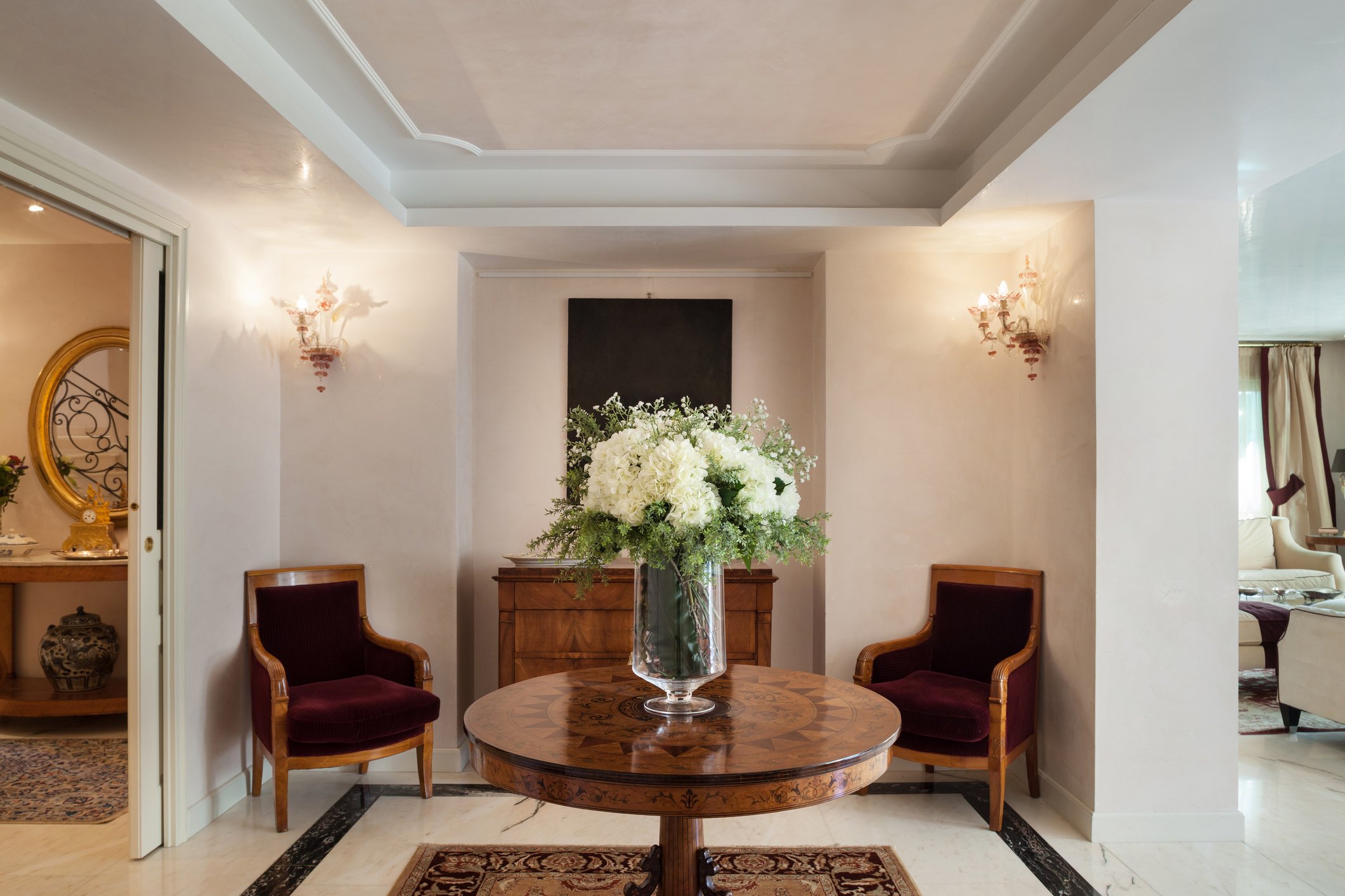 Elegant room with a round wooden table featuring a large bouquet of white flowers, two burgundy armchairs, wall sconces, and a mirror on the left wall.