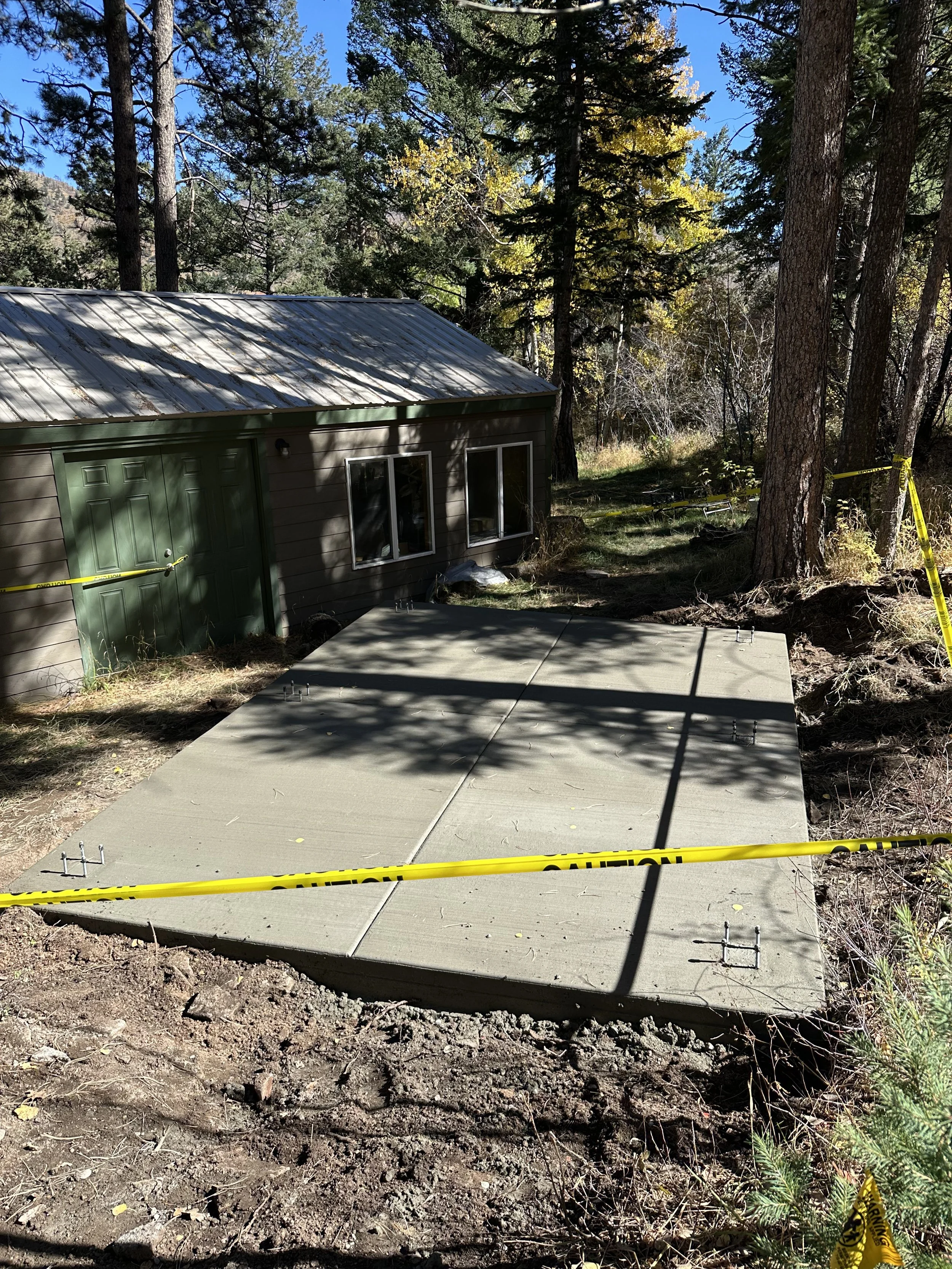 Photo of a freshly poured concrete patio in a wooded area with large trees. The surrounding area is marked off with yellow caution tape.