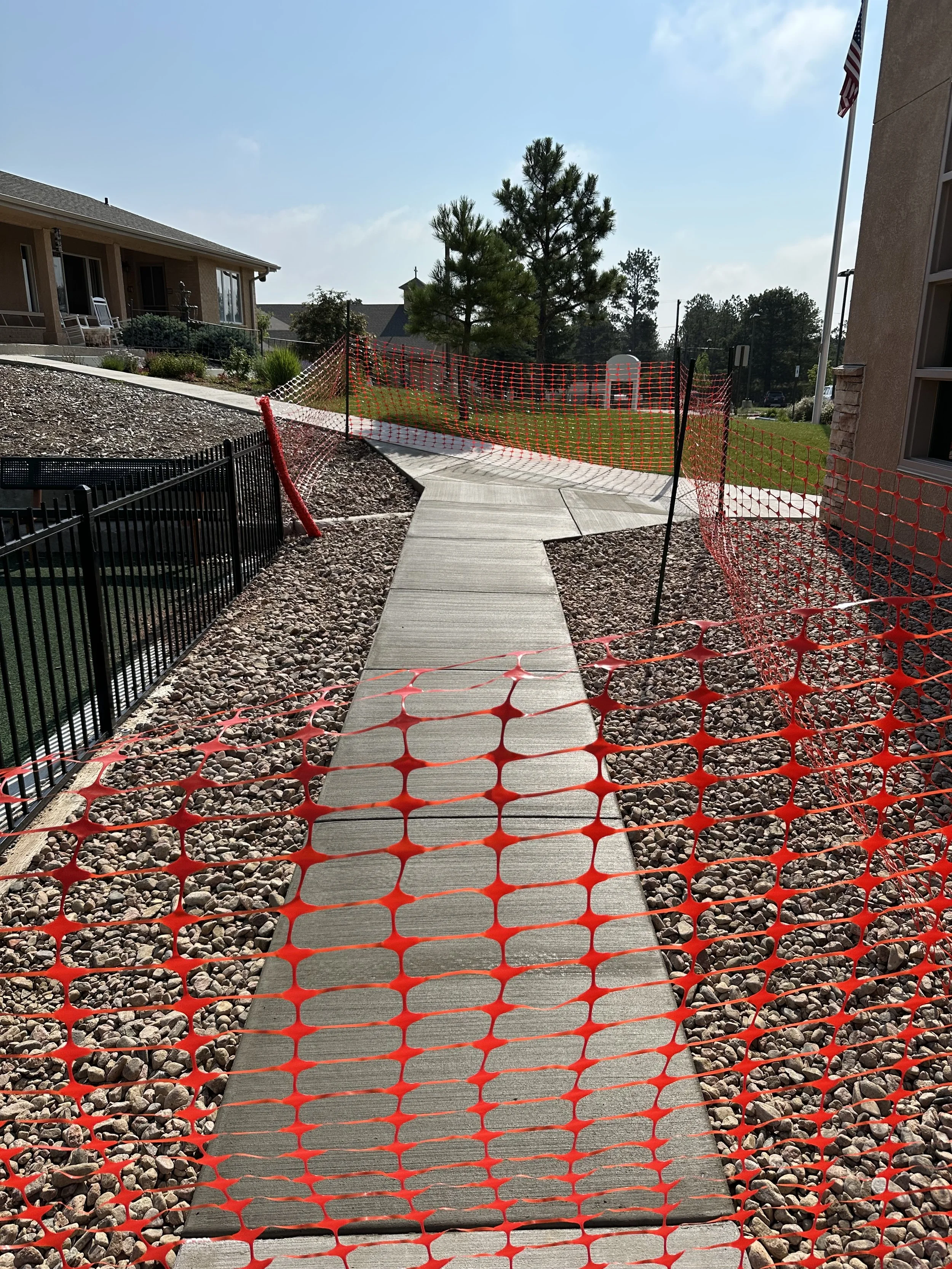 Sidewalk construction site with orange safety fencing, a concrete pathway, gravel, a few buildings, trees, and a blue sky.