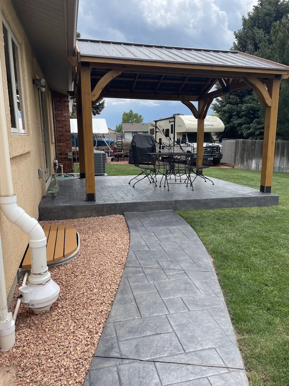 Backyard patio with a wooden gazebo, metal outdoor furniture, a recreational vehicle, a boat, and a grassy yard with a wooden fence.