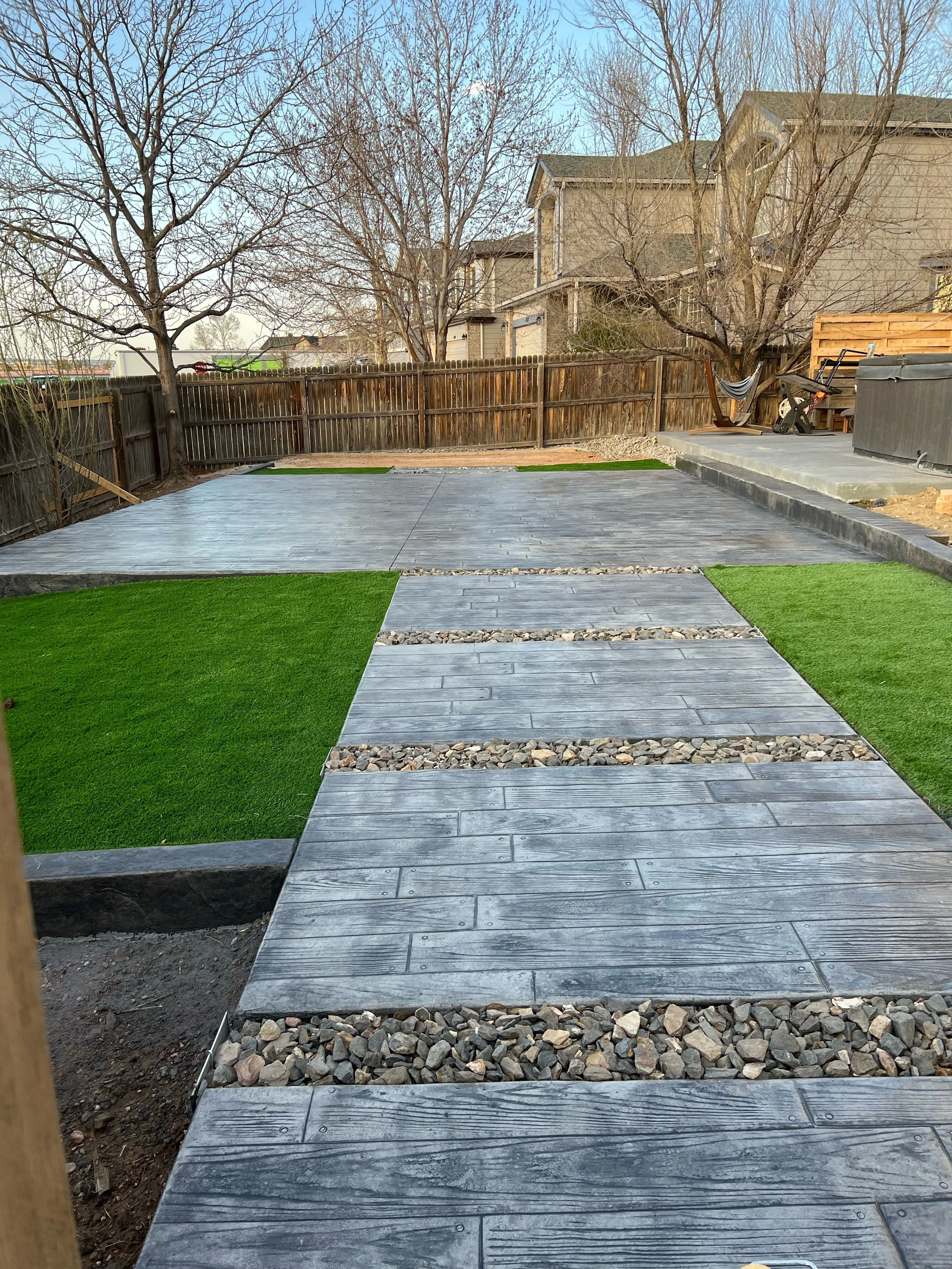 Backyard patio with gray wooden tiles, green artificial grass patches, and a fence with trees in the background.