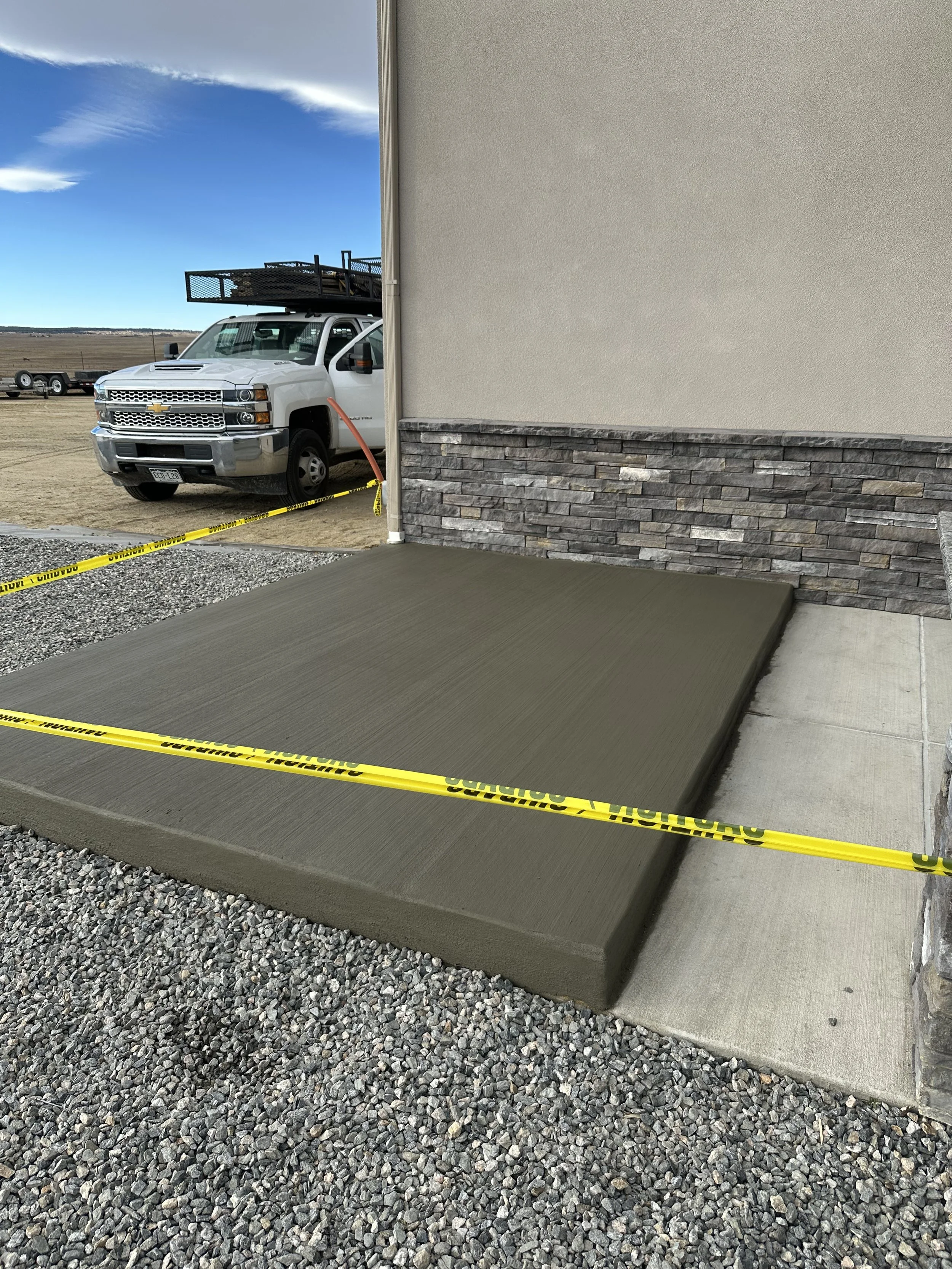 Construction area with a freshly poured concrete slab, yellow caution tape, gravel ground, a white pickup truck with a roof rack in the background, and a building wall with stone veneer and beige stucco.
