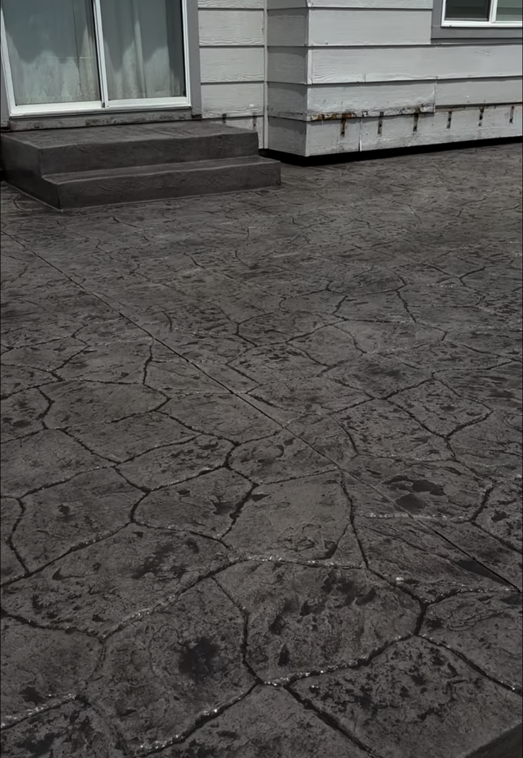 A close-up view of a concrete patio with textured paving stones in front of a house. The house has a sliding glass door with curtains and gray wooden siding with a small window. There are steps leading up to the door and some rusted metal brackets on