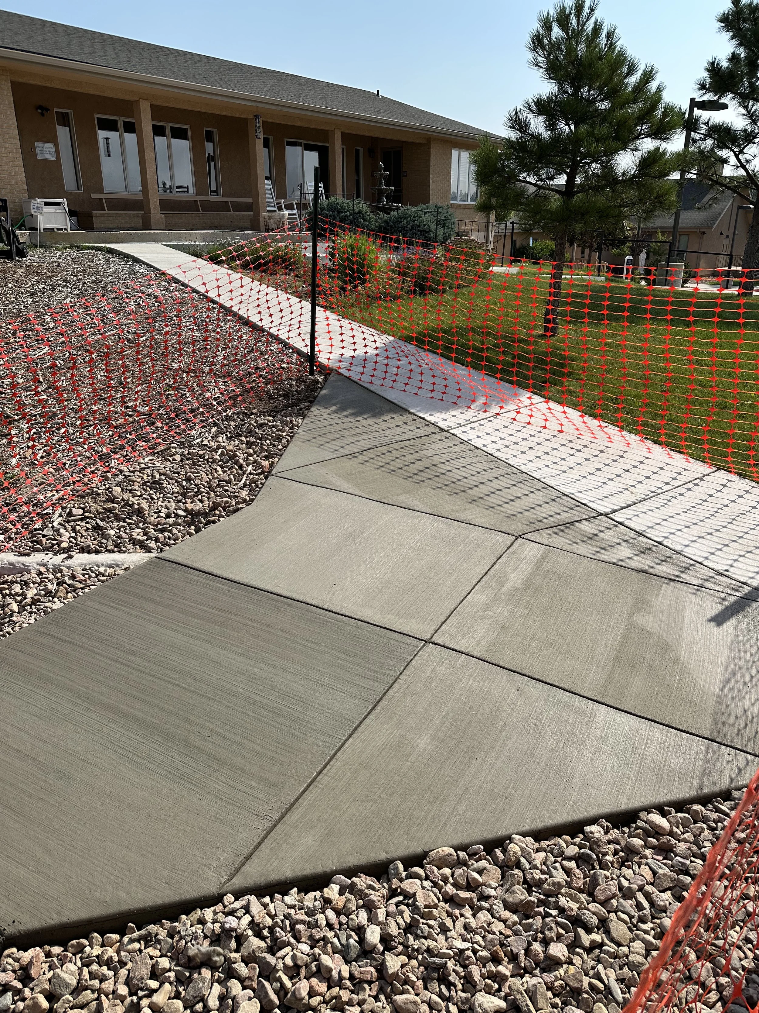 New concrete sidewalk being installed next to a house with a gravel yard, surrounded by orange safety fencing, with trees and neighboring houses in the background.