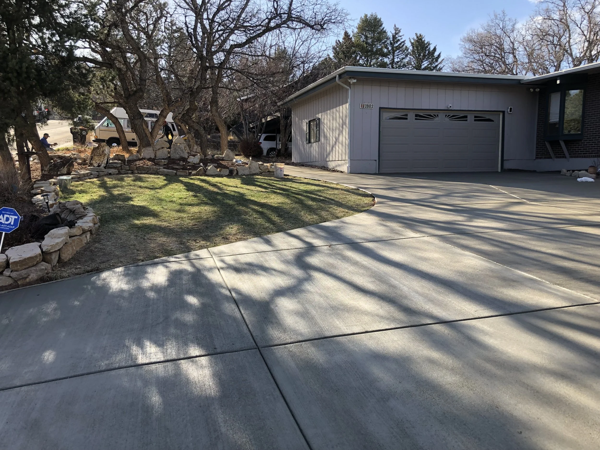 Residential driveway leading to a two-car garage with a light gray door. To the left, a small lawn with a stone border and leafless trees casting shadows. There is a security sign near the stone border and several vehicles parked behind trees on the left side of the image.