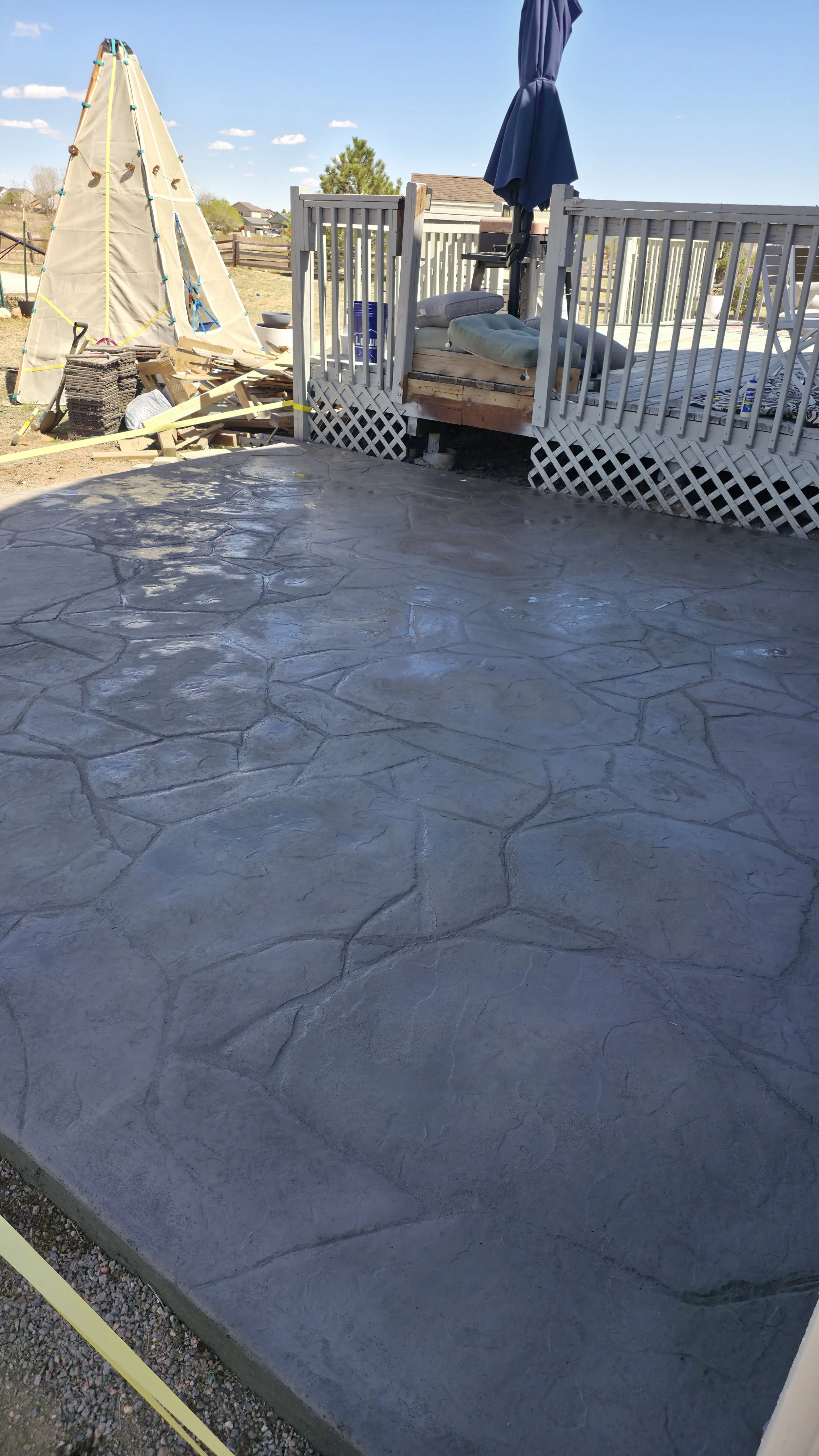 Concrete patio with stamped pattern, part of a backyard with a wooden deck, umbrella, and a backyard landscape with houses and trees in the background.