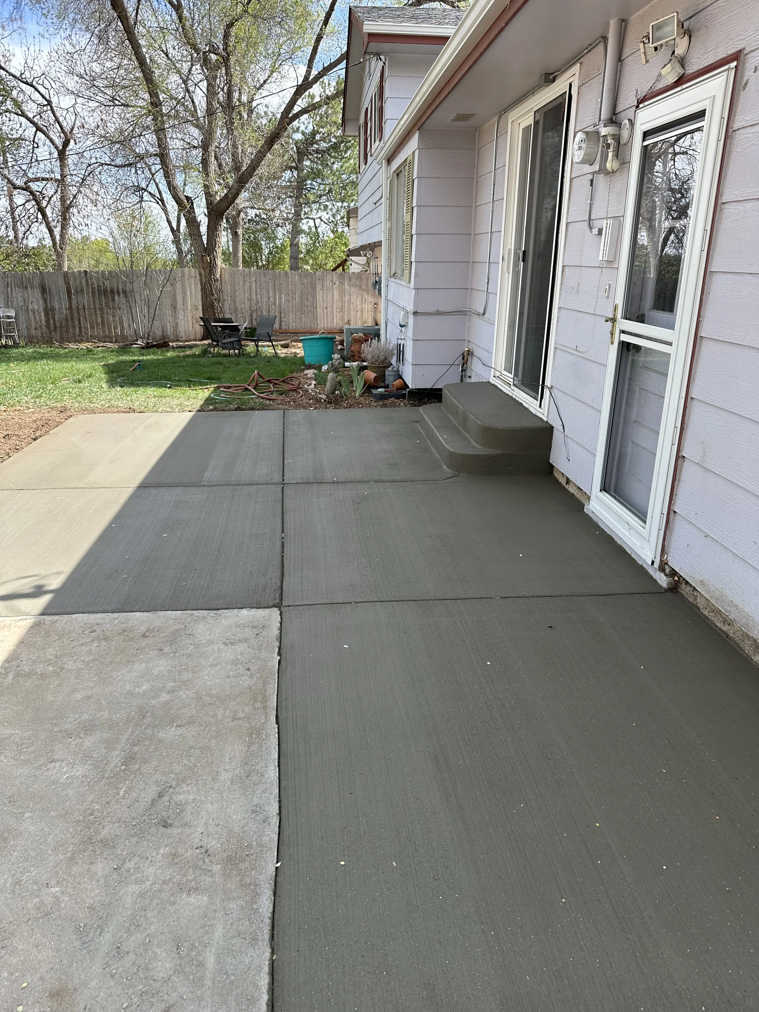 A backyard patio with newly poured concrete slabs leading to a grassy yard with a patio table and chairs, surrounded by a wooden fence and trees.