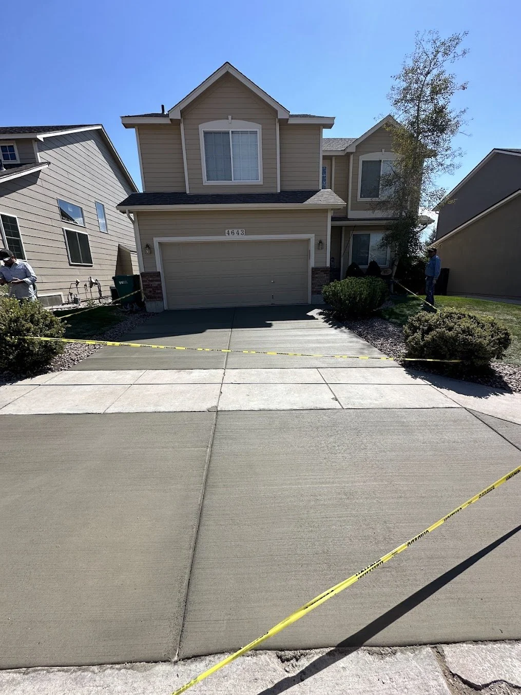 Newly paved concrete driveway in front of a beige two-story house with a closed garage door, surrounded by caution tape, with two workers on either side, and trees and neighboring houses in the background.
