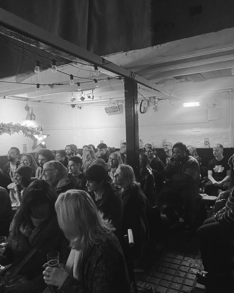 A large group of people seated indoors at a concert or event, with some holding drinks. The room has string lights hanging from the ceiling and a star-shaped decoration with a light on the wall.