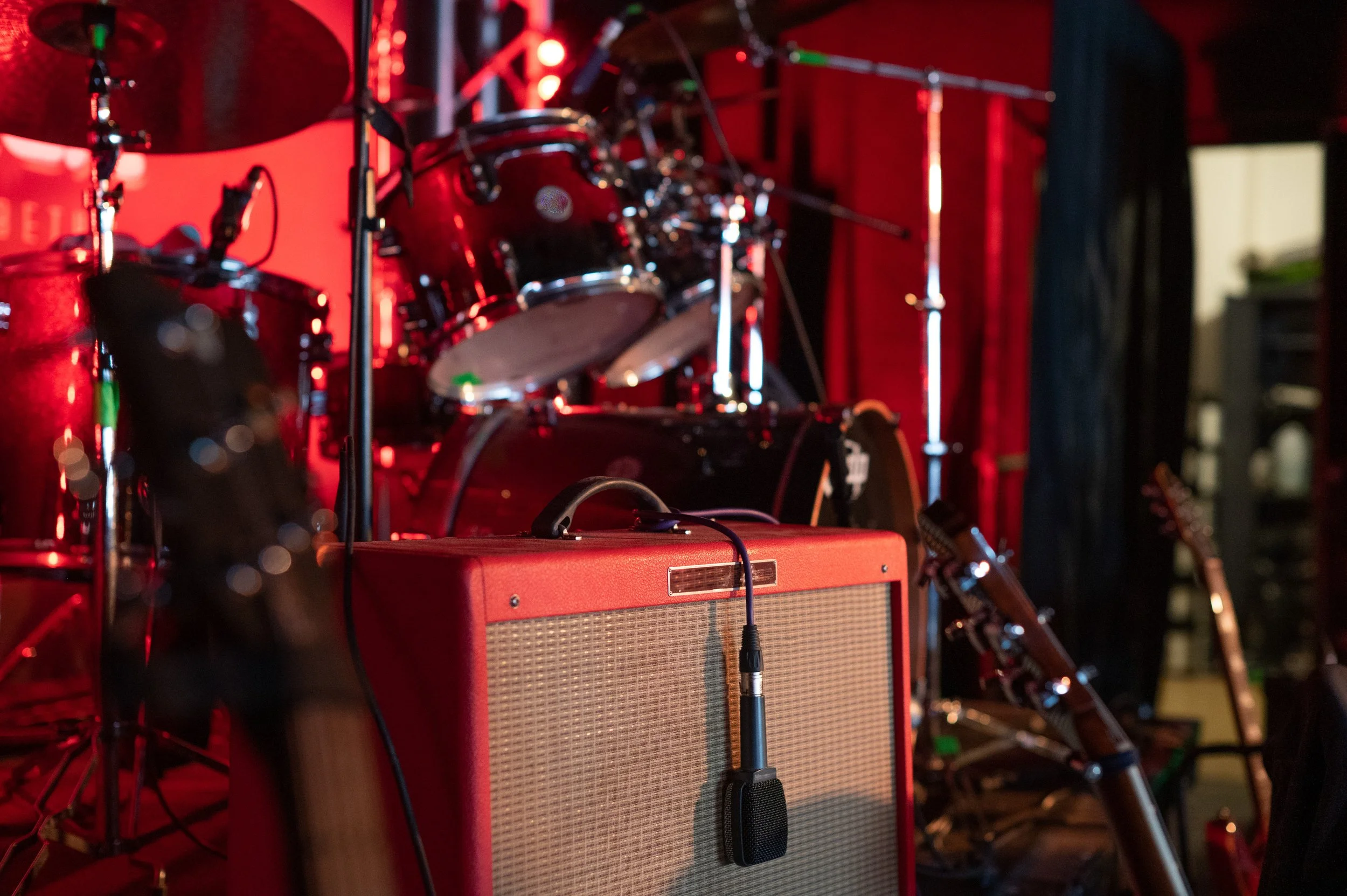 A red amplifier with a microphone on it, electric guitars, drums, and microphones in a music stage setup with red lighting and a drum set in the background.