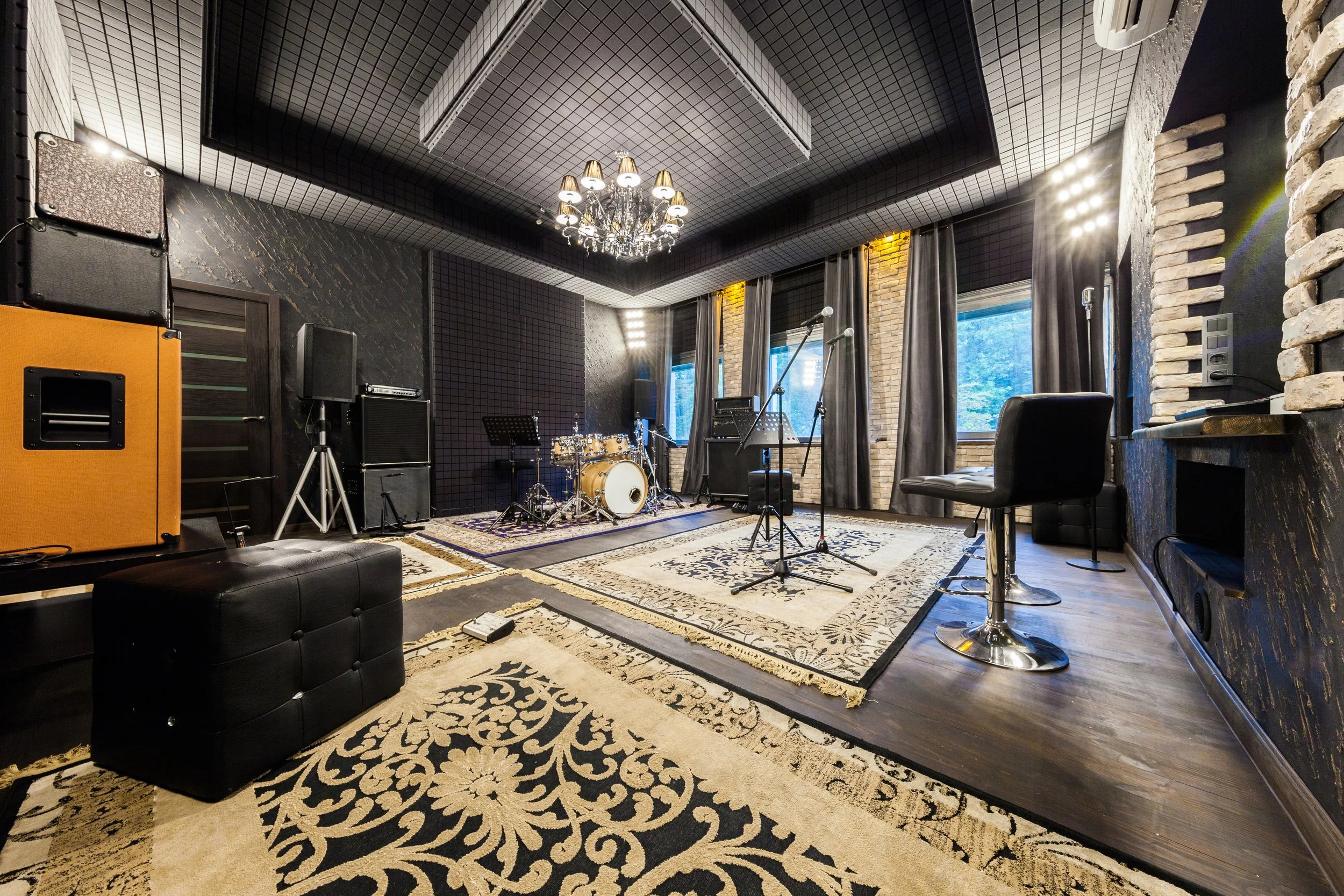 Music recording studio with drums, microphones, amplifiers, and seating, featuring black and gold decor with patterned rugs and large windows.