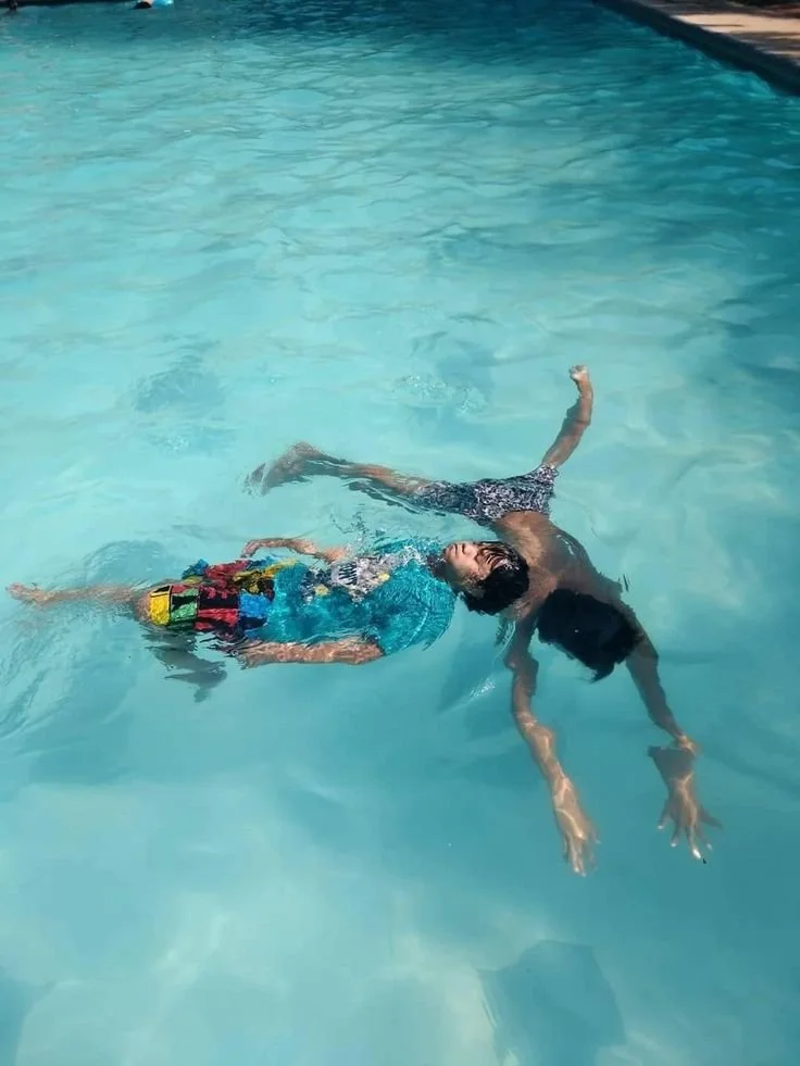 Two children are floating and playing in a swimming pool. One child is on their back smiling, while the other child is face down reaching towards the water.