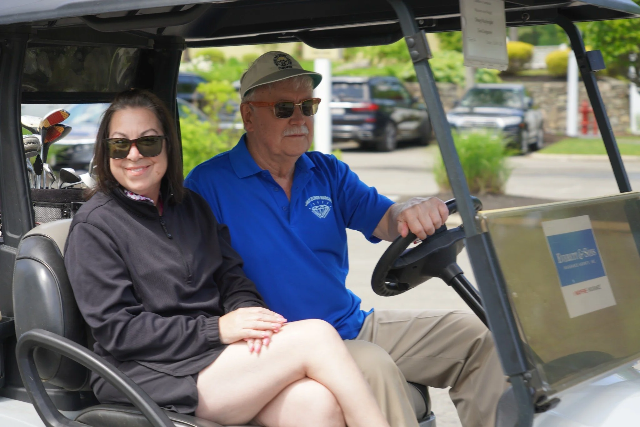 A woman and a man sitting in a golf cart, with the woman smiling and wearing sunglasses and the man driving, outdoors in a parking lot.