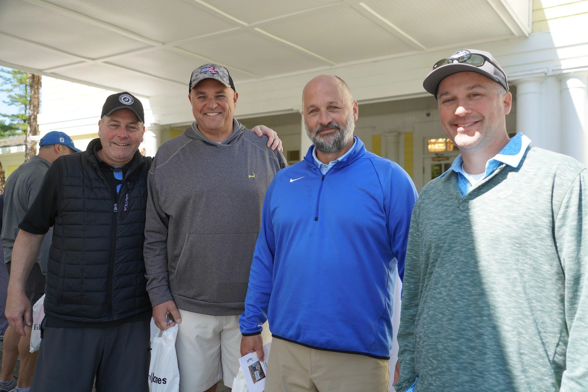 Four men standing together outdoors, smiling, in a covered porch area. They are dressed casually, with some wearing golf caps and sunglasses.