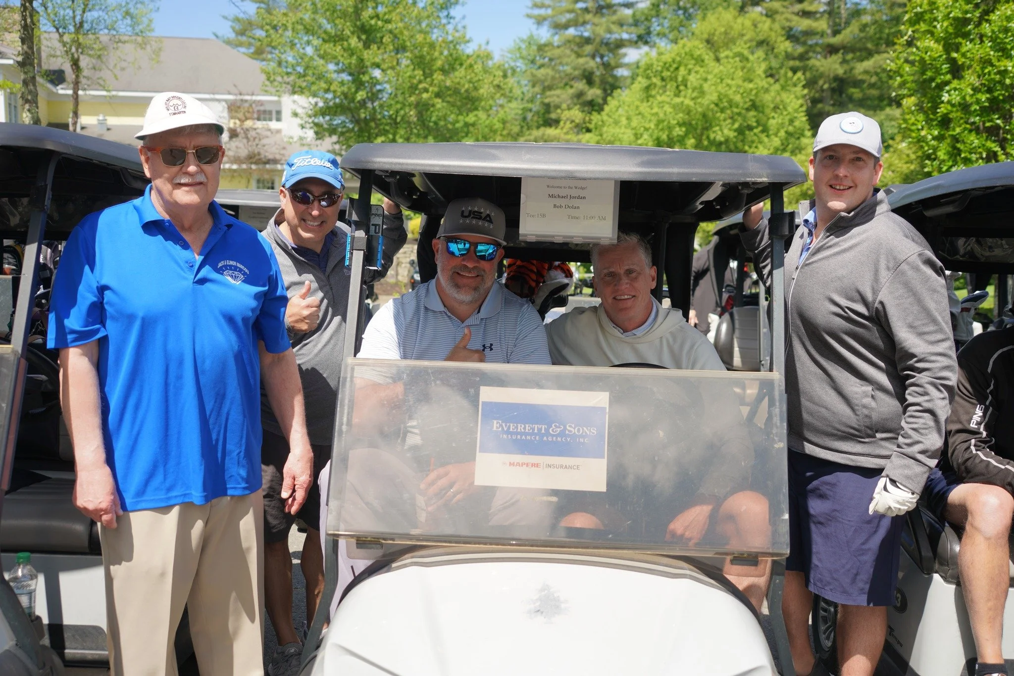 Group of five men posing around a golf cart at a golf course, with green trees and clear sky in the background.